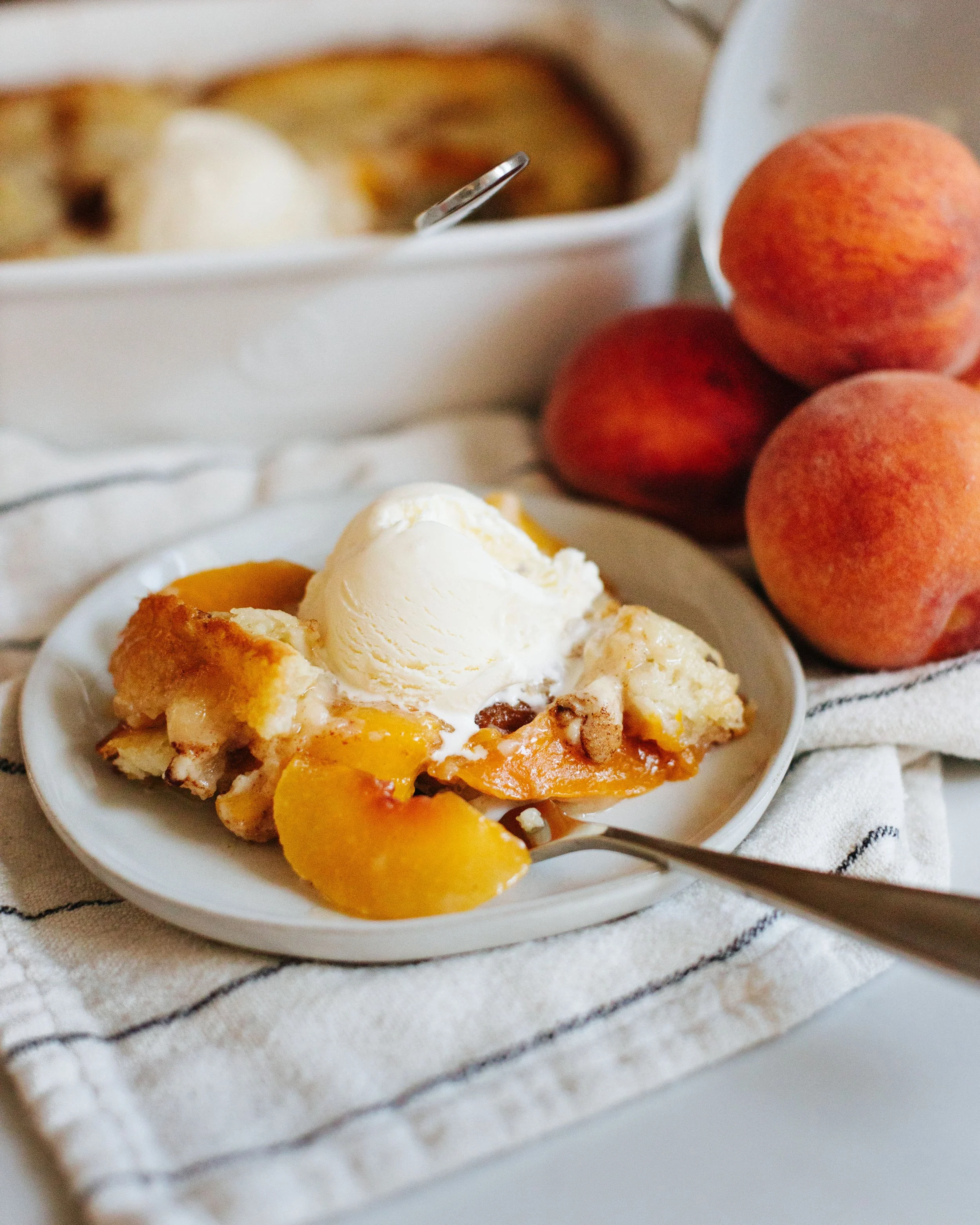 Peach cobbler with a scoop of vanilla ice cream on top, served on a white plate with fresh peaches nearby.