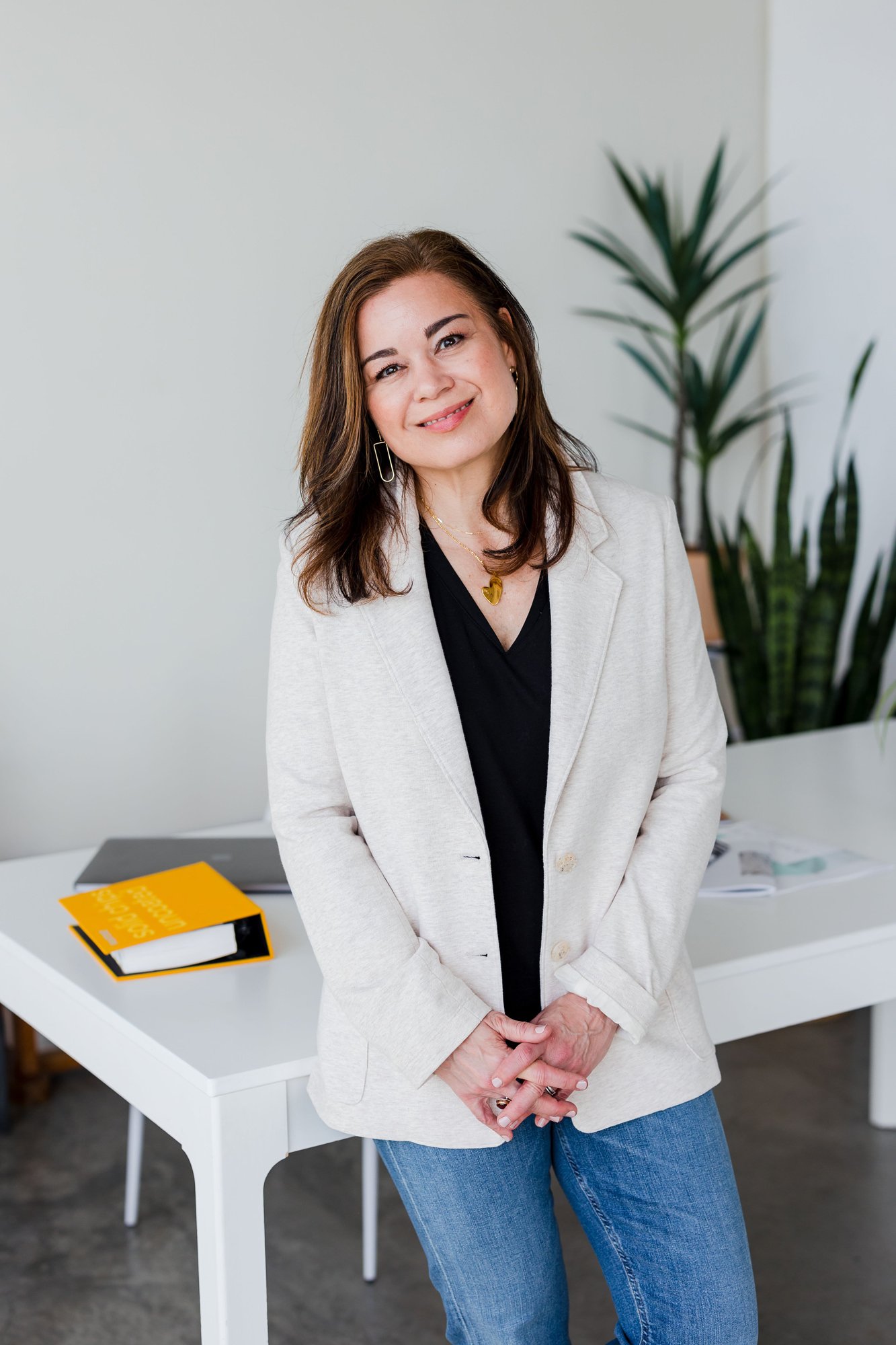 A woman with shoulder-length brown hair, wearing a cream blazer over a black top, and blue jeans, standing in an office space with a large green plant in the background and a white desk with a yellow notebook and some papers.