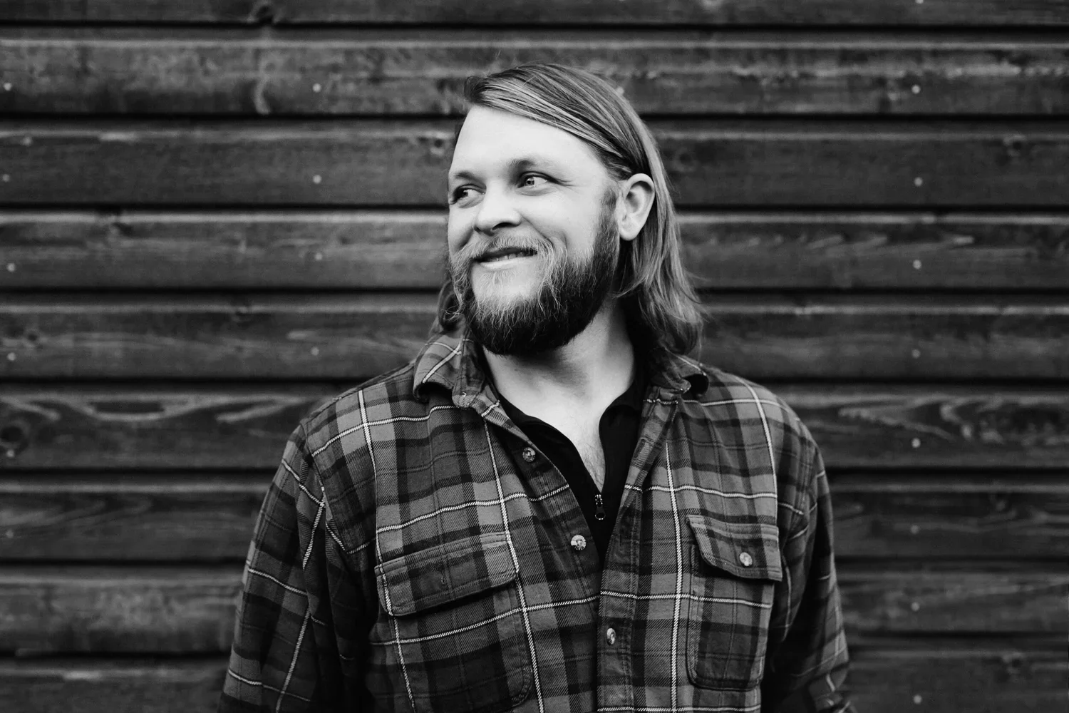 A man with long hair and a beard, wearing a plaid shirt, smiling and looking to the side against a wooden background.