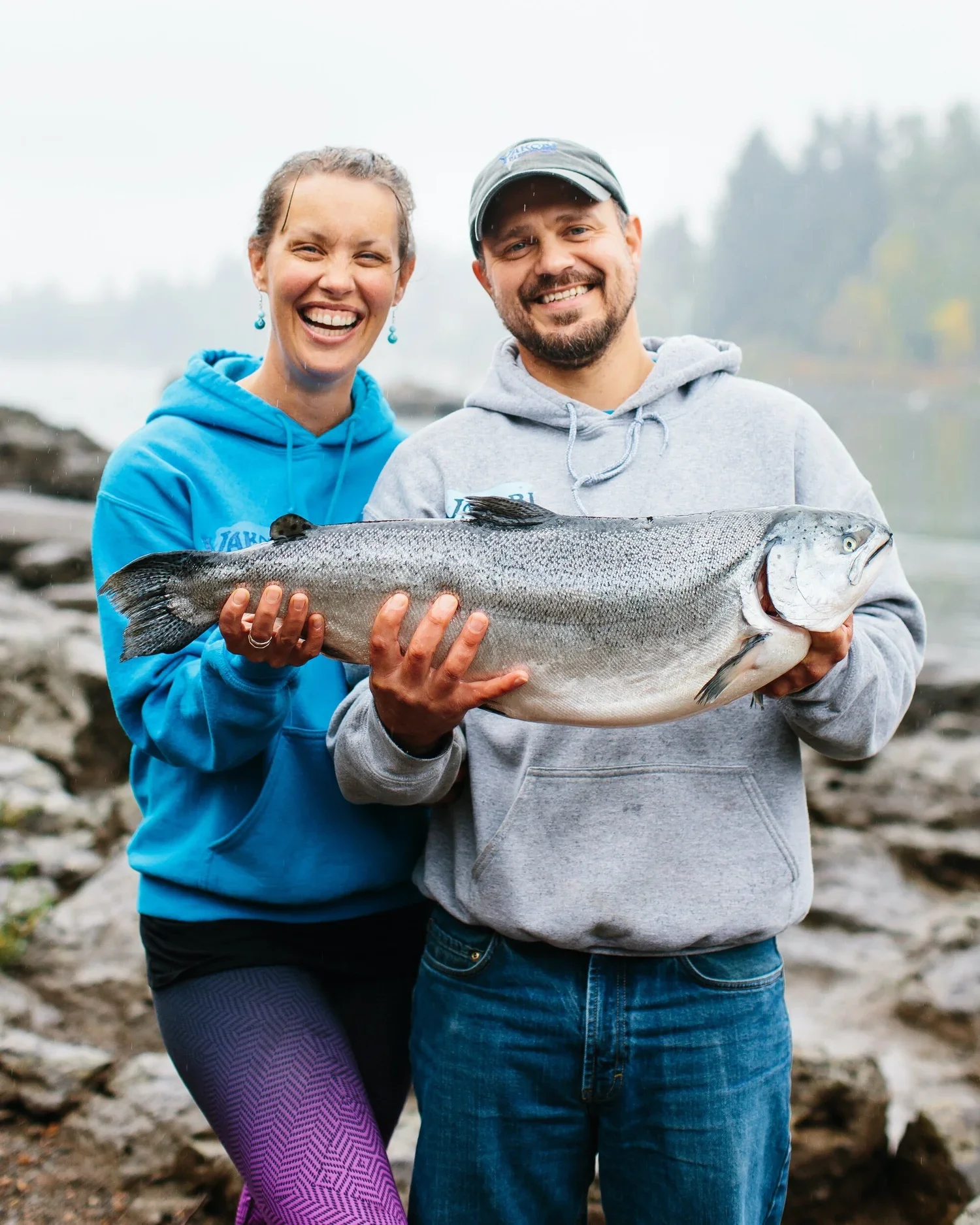 A smiling man and woman holding a large fish outdoors near water. They are dressed in casual hoodies, with a rocky shoreline and trees in the background.