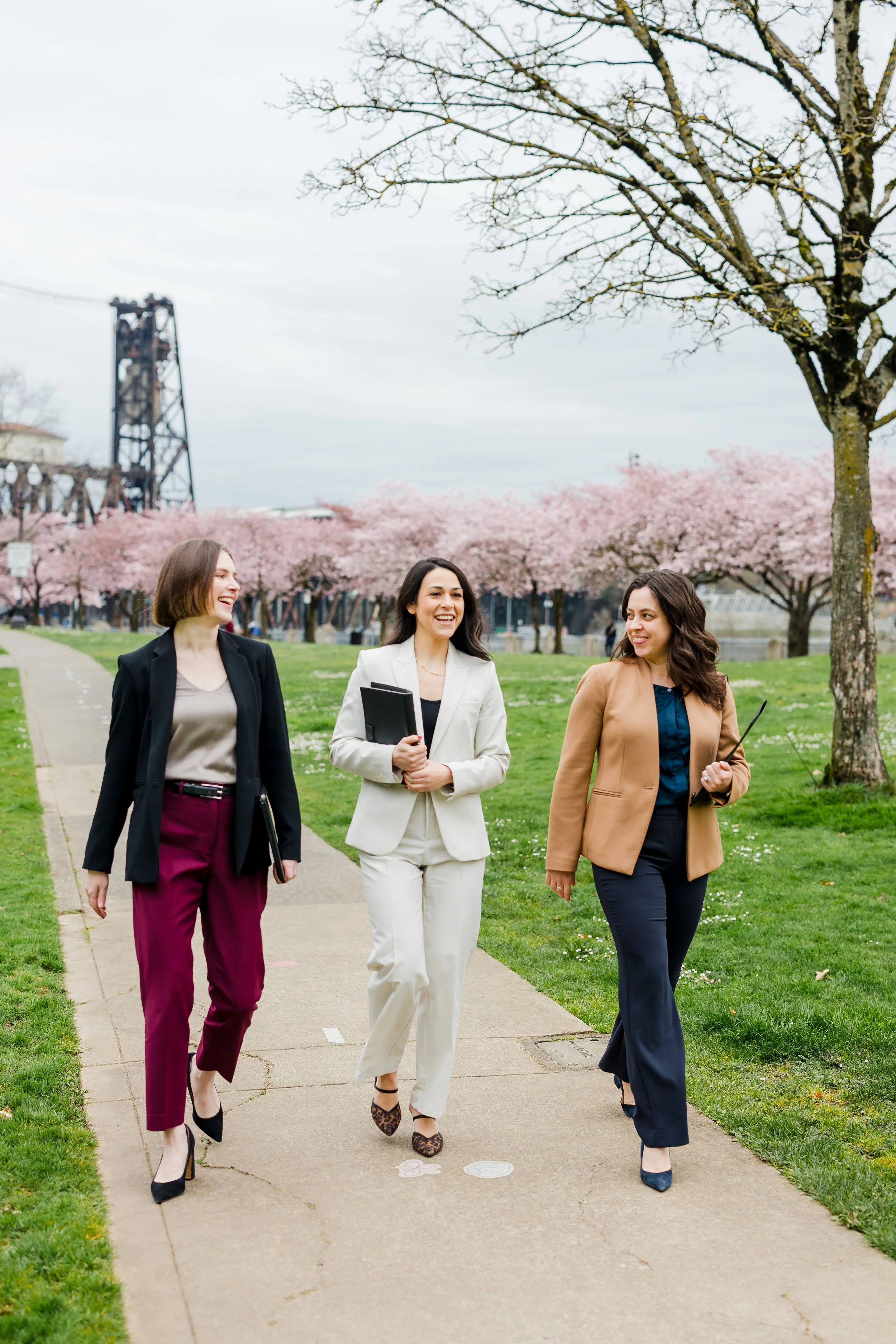 Three women walking and talking outdoors on a sidewalk, surrounded by blooming cherry blossom trees, with a cloudy sky and a distant industrial structure.