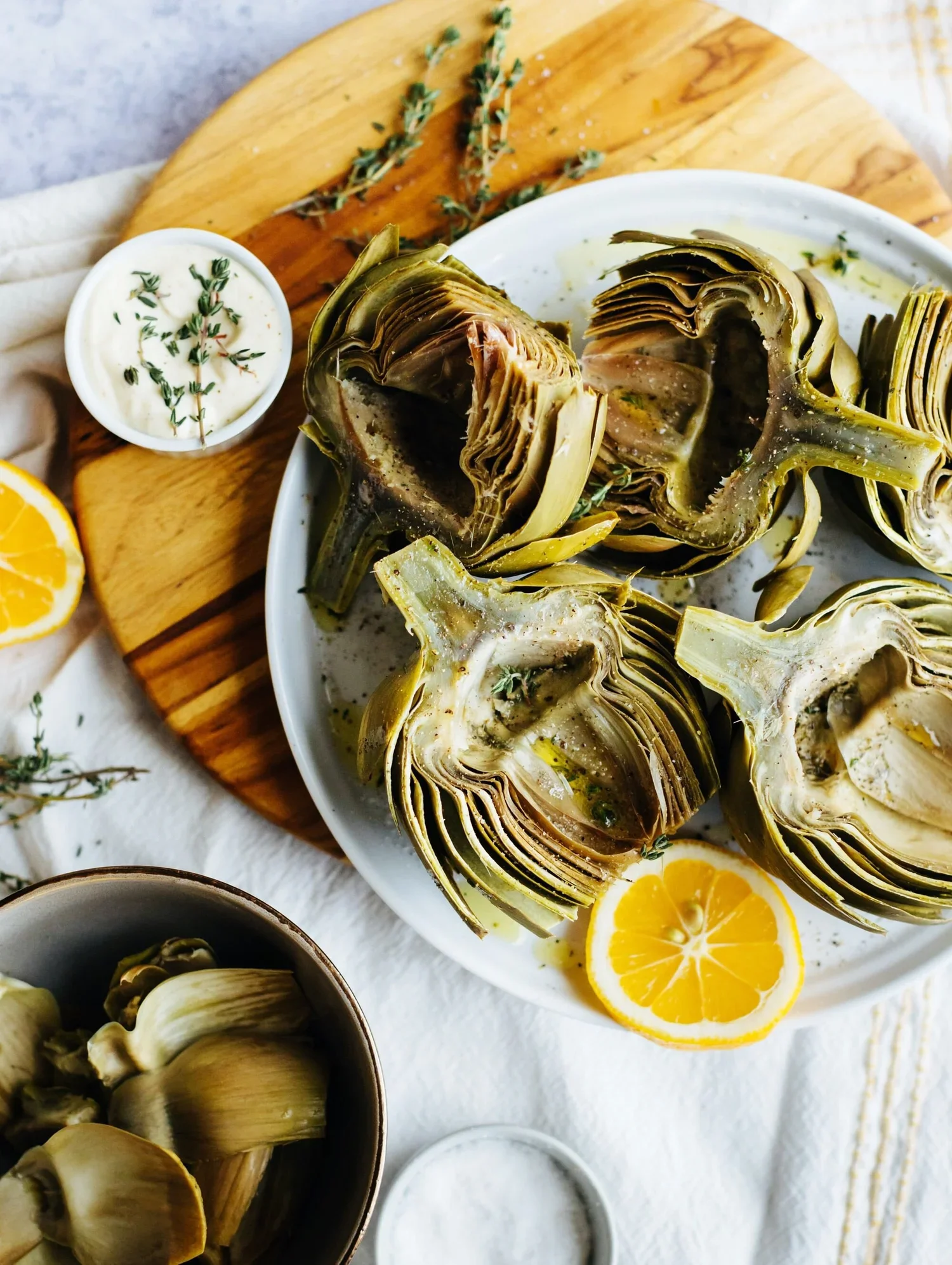 A plate of cooked artichokes garnished with lemon slices and herbs, with a side of dipping sauce, on a wooden cutting board with a small bowl of artichoke hearts and a lemon half nearby.