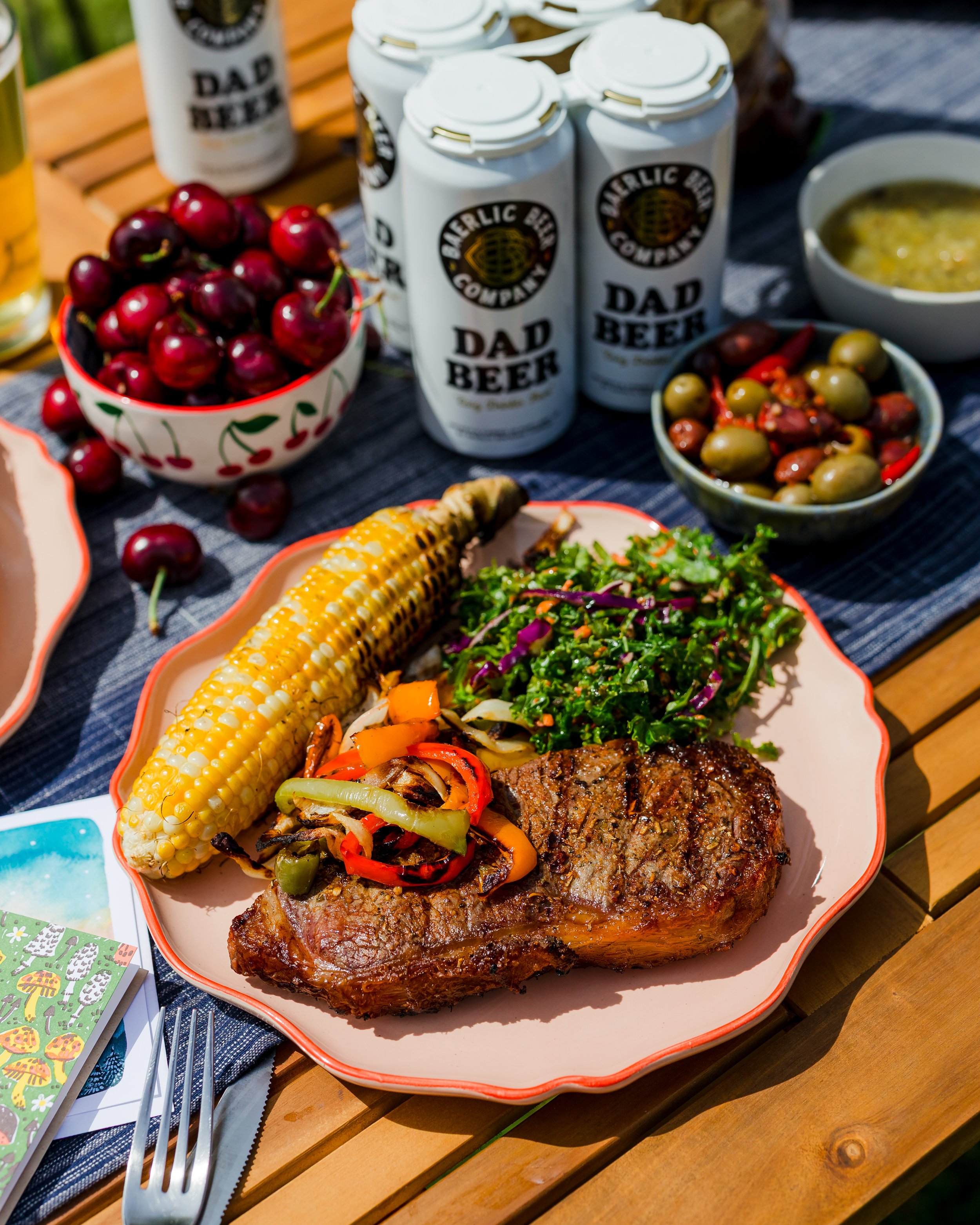 A plate of grilled steak with grilled vegetables, a cob of corn, and a side salad, surrounded by bowls of cherries and olives, cans of beer, and other food items on a wooden outdoor table.
