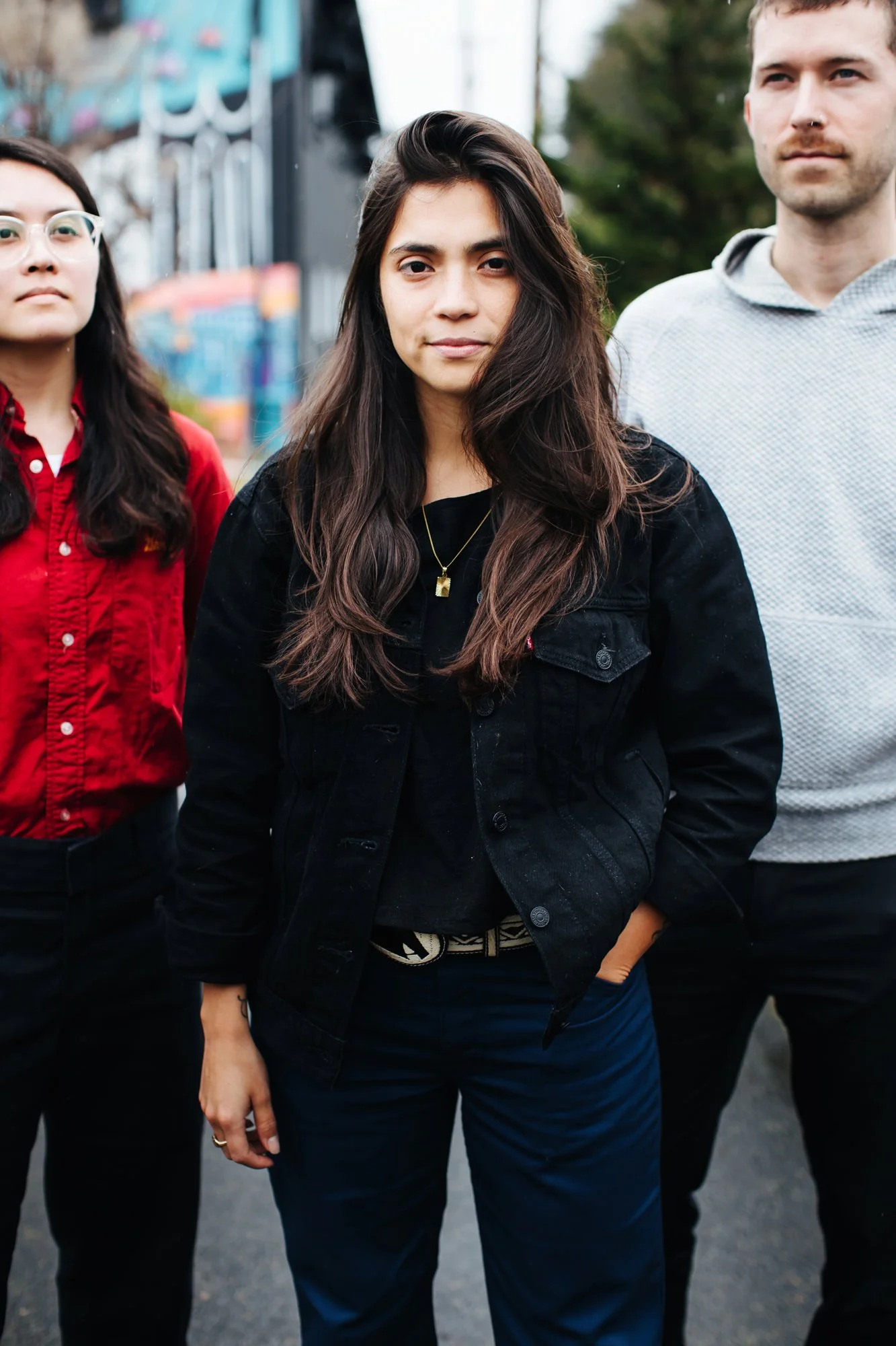 Three young adults standing outdoors on a street, with a mural in the background, one woman and two men, wearing casual clothes.