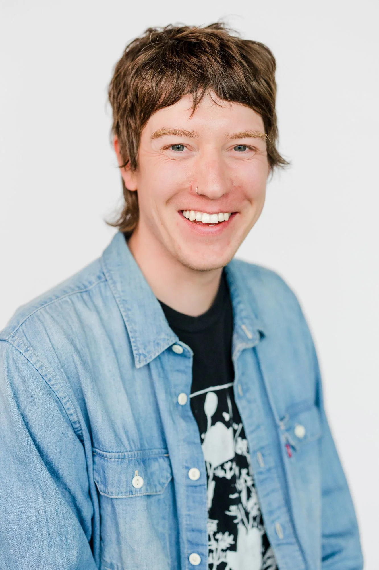 A young man with brown hair, blue eyes, and a nose piercing, smiling and wearing a black t-shirt with a graphic design and a light blue denim shirt, against a plain white background.