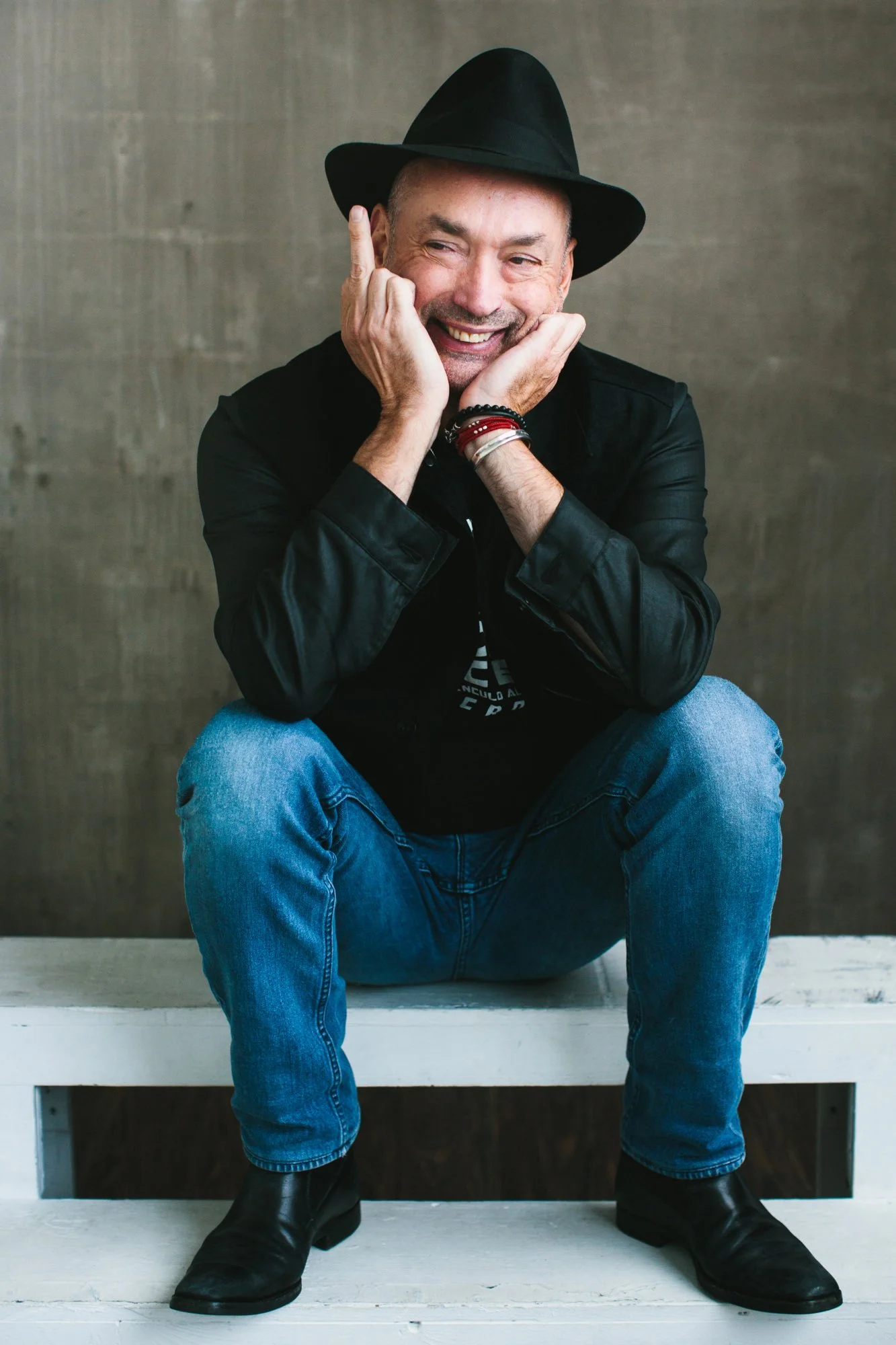 Man wearing a black hat, black jacket, and jeans sitting on a white bench with a gray background, smiling and posing with his hands near his face.