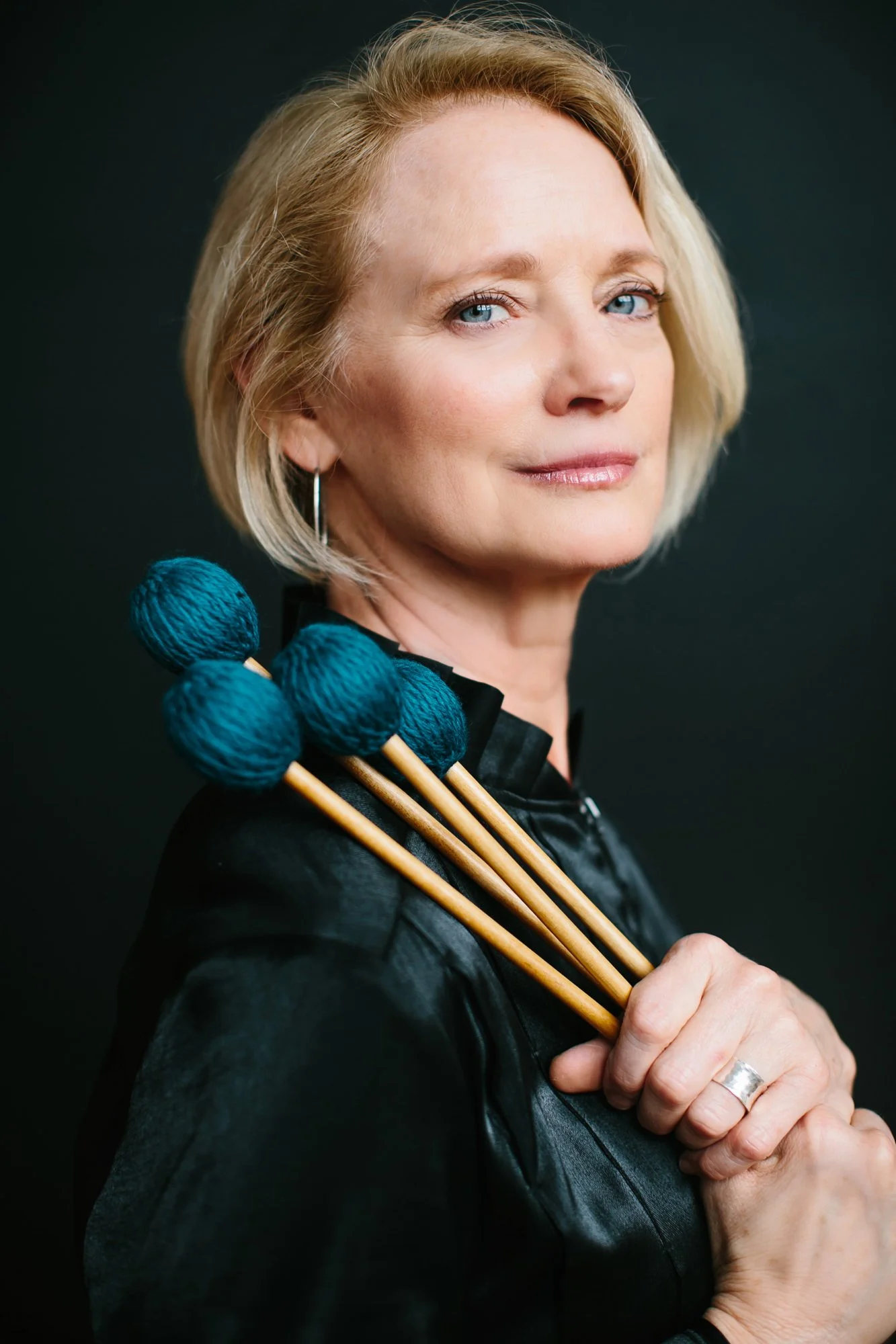 A woman with blonde hair and blue eyes holding four teal knitting needles with yarn balls attached to them, wearing a black top, standing against a dark background.
