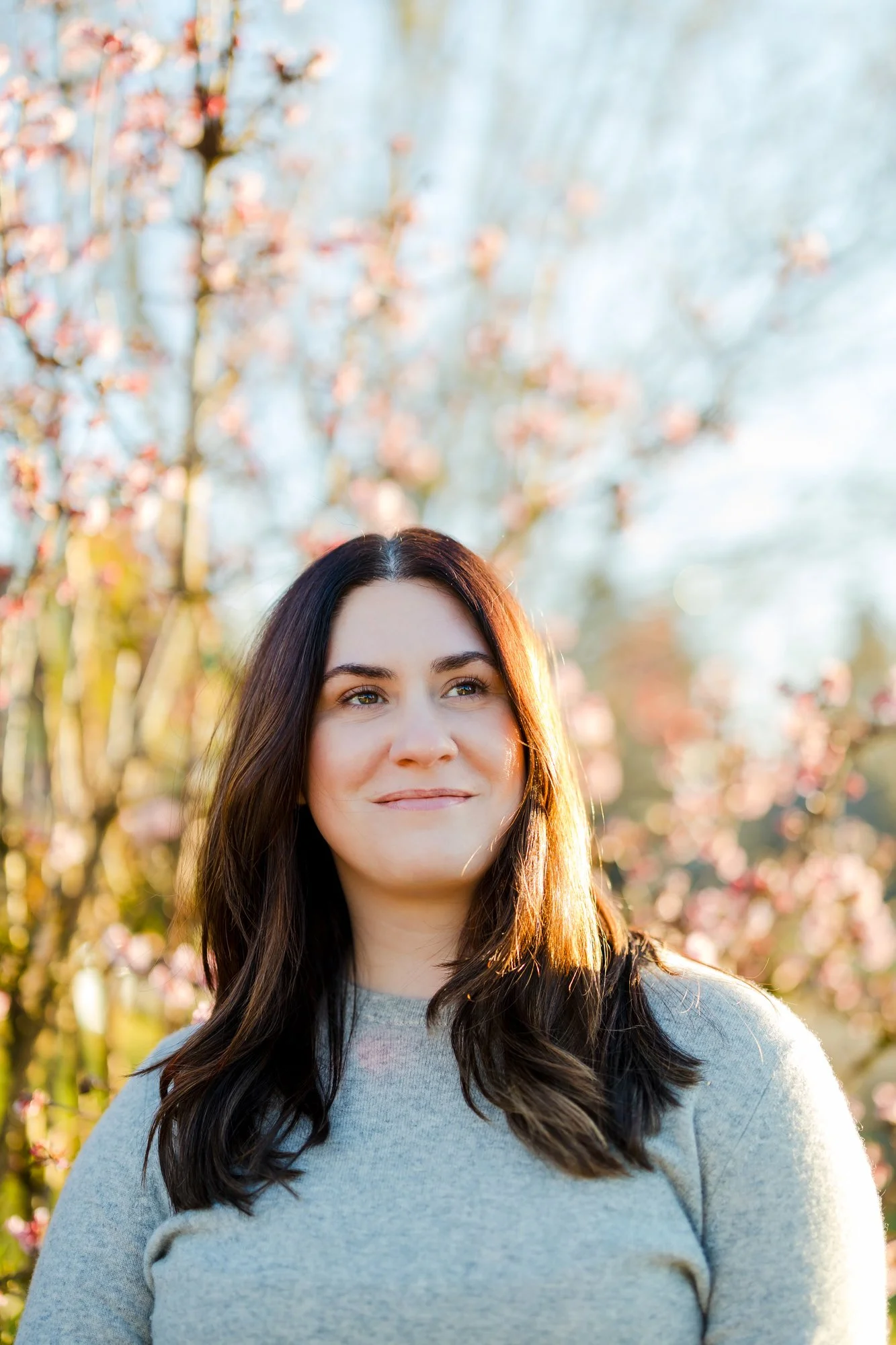 A woman with dark brown hair and light skin standing outdoors near blooming pink flowers with sunlight illuminating her face.