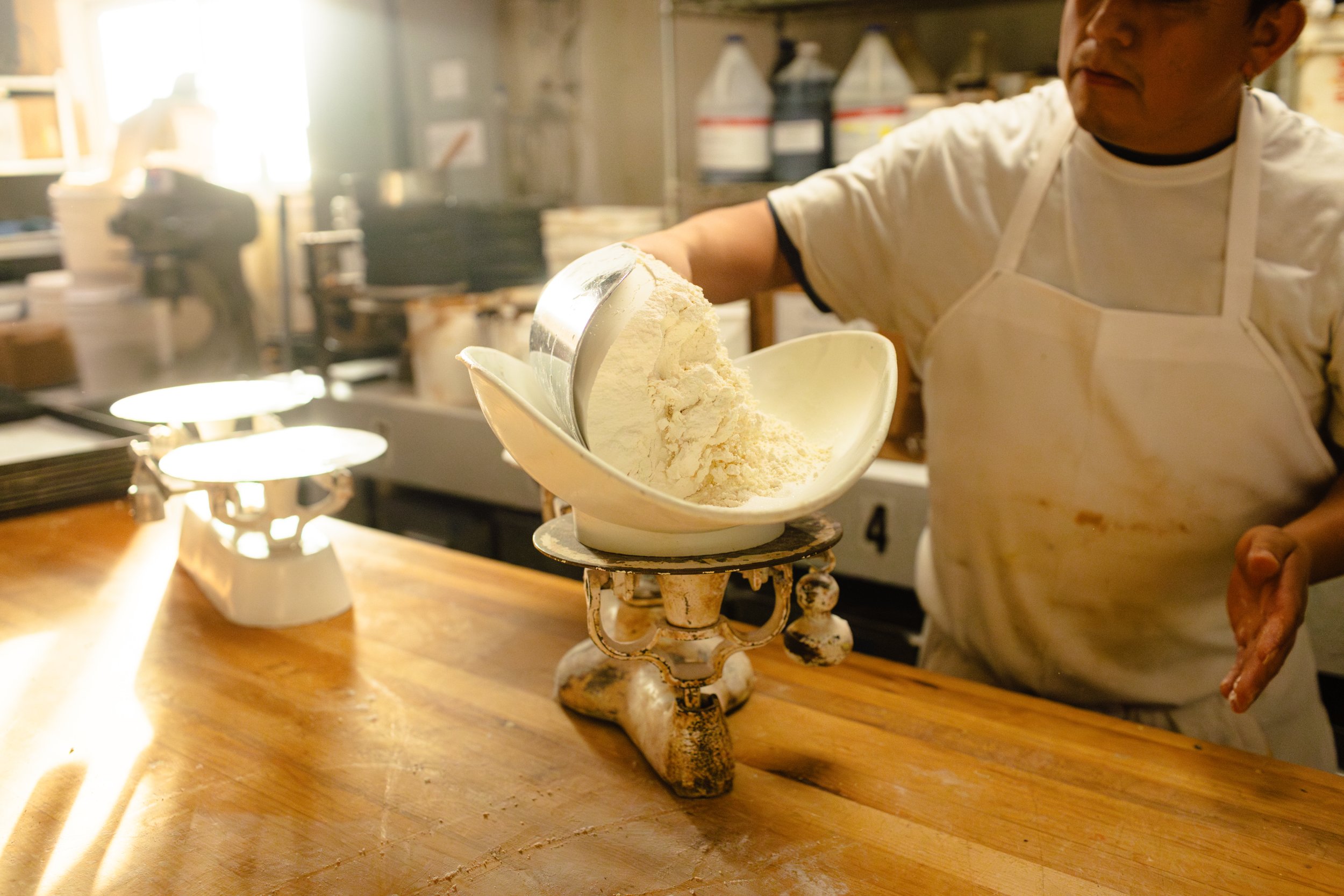 A person in a white apron is pouring flour from a metal scoop into a bowl on a vintage scale in a kitchen.