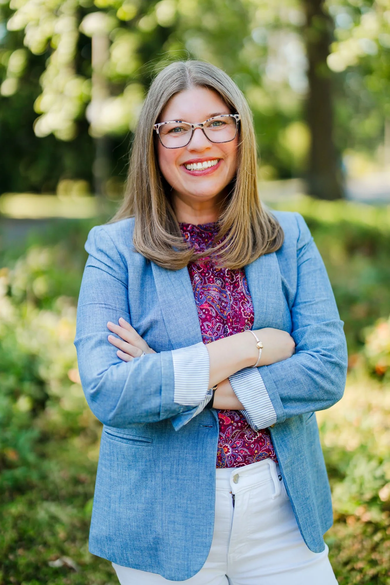 A smiling woman with shoulder-length light brown hair, wearing glasses, a blue blazer over a red patterned blouse, and white pants, standing outdoors in a park with green trees and grass in the background.