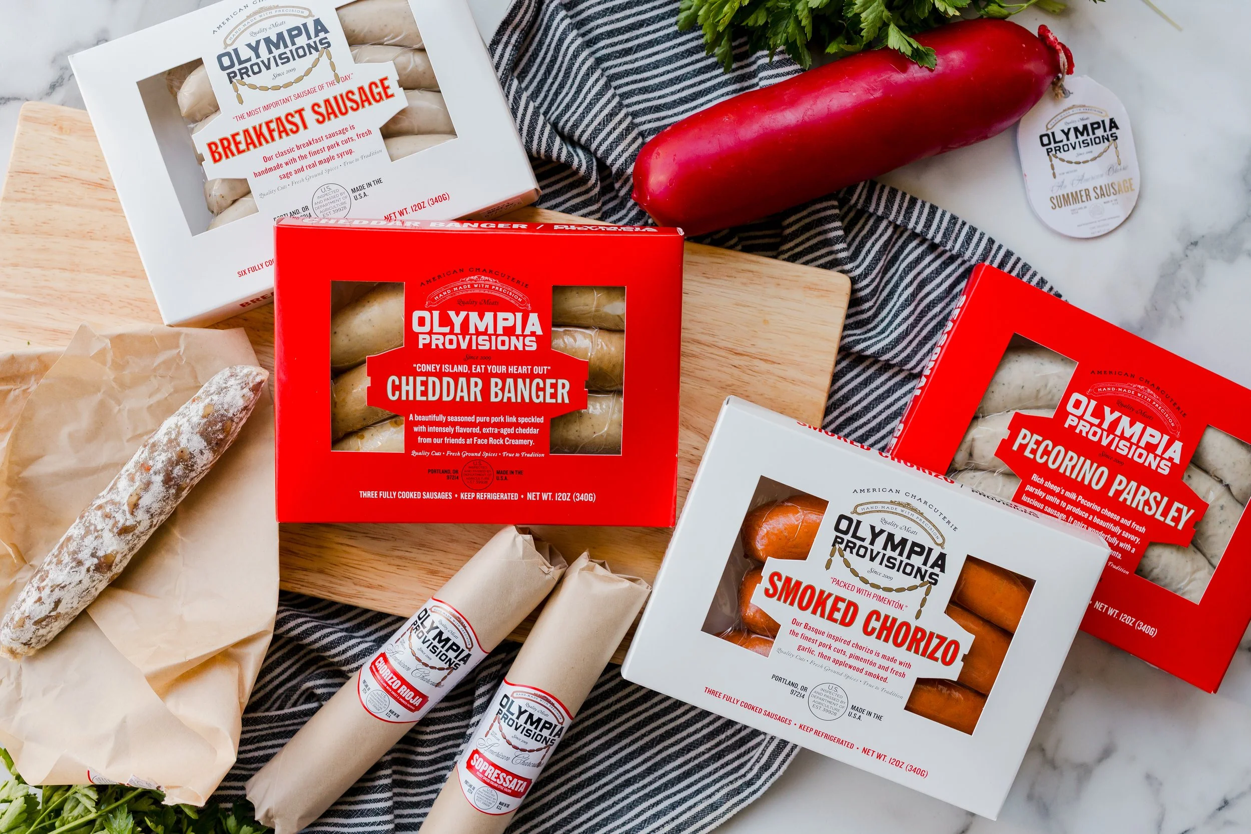 Assorted Olympia Provisions sausages and meats, including breakfast sausage, cheddar banger, pecorino parsley, and smoked chorizo, displayed on a wooden surface with a red radish, green herb, and a striped cloth.
