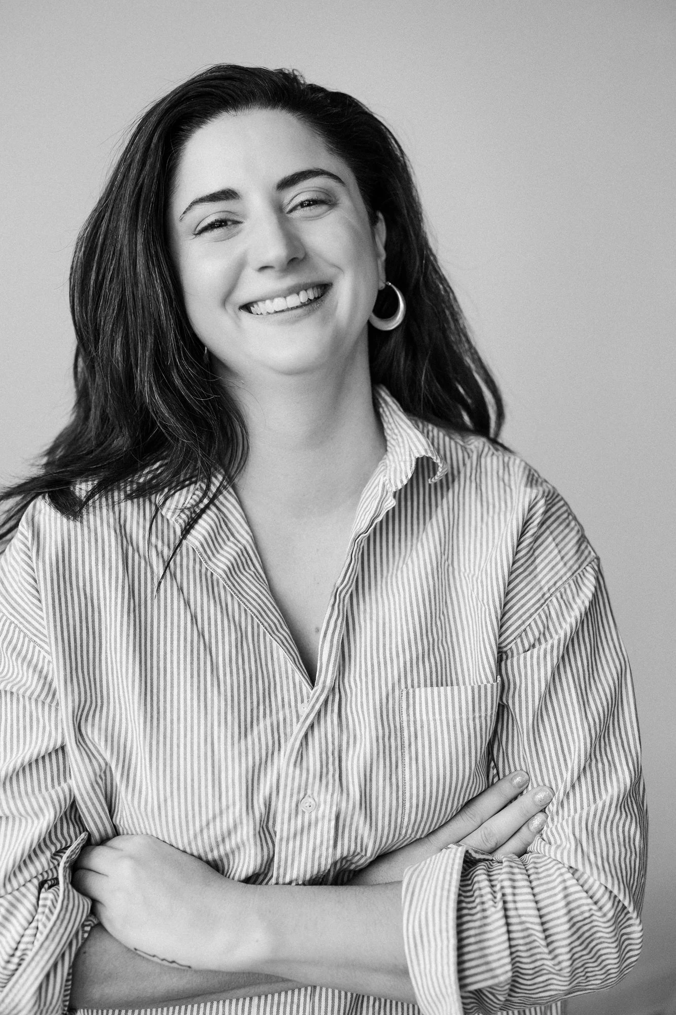 A smiling woman with dark hair, wearing a striped shirt and hoop earrings, standing with arms crossed against a plain light background.