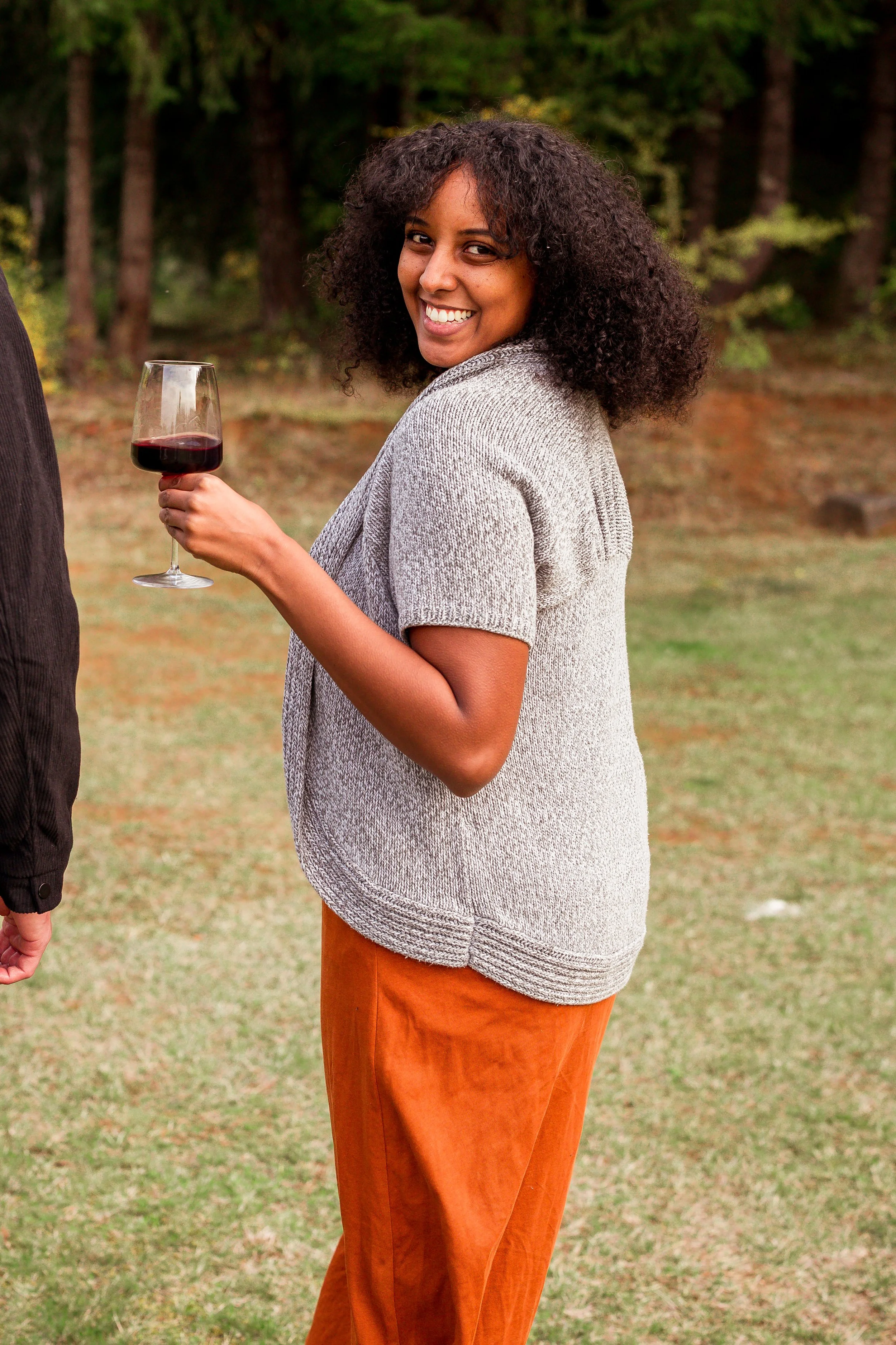 A woman with curly hair smiling and holding a glass of red wine outdoors, with trees in the background.