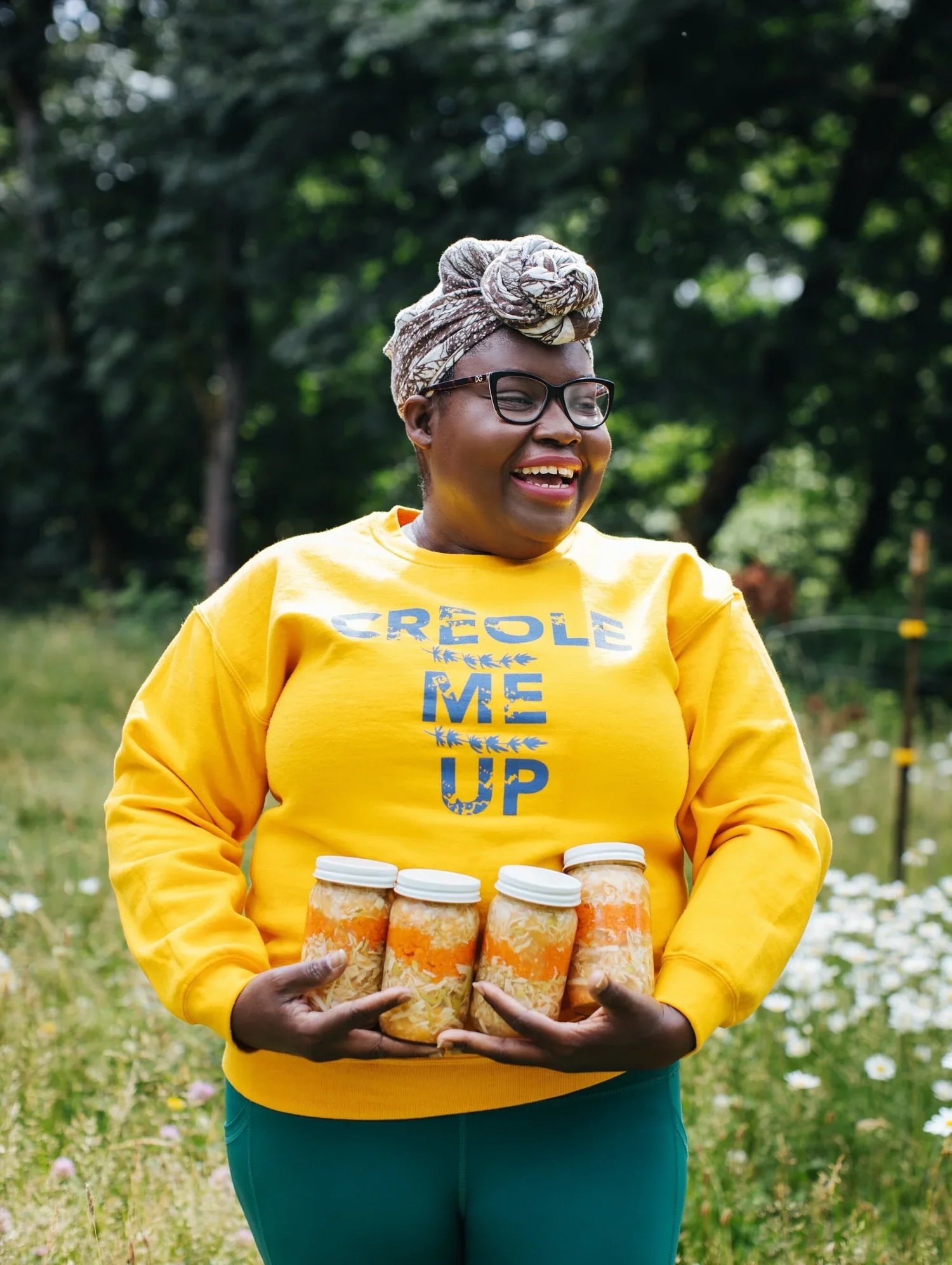 A woman smiling outdoors, wearing a yellow sweatshirt with blue text, a headwrap, and glasses, holding four jars in her hands.