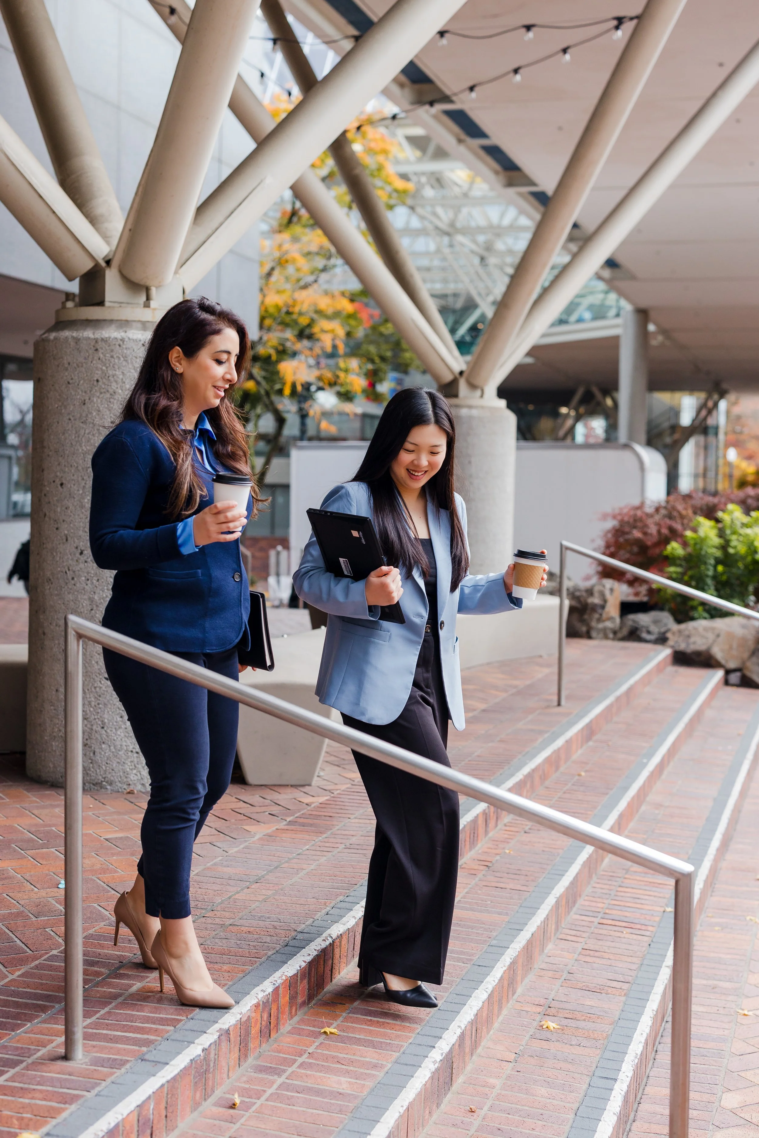 Two women walking down outdoor stairs, each holding a coffee cup and smiling, with a modern architectural building and autumn foliage in the background.