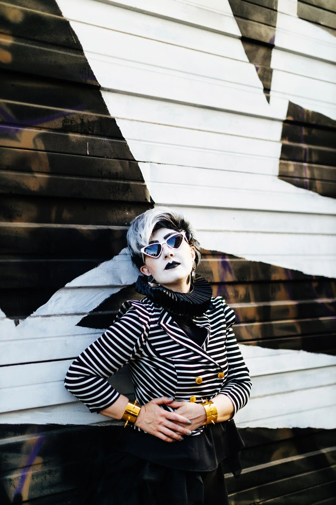 A woman with black lipstick, wearing purple sunglasses, a black and white striped blazer with gold buttons, and gold cuffs on her wrists, poses in front of a graffiti-covered wall with black, white, and brown colors.