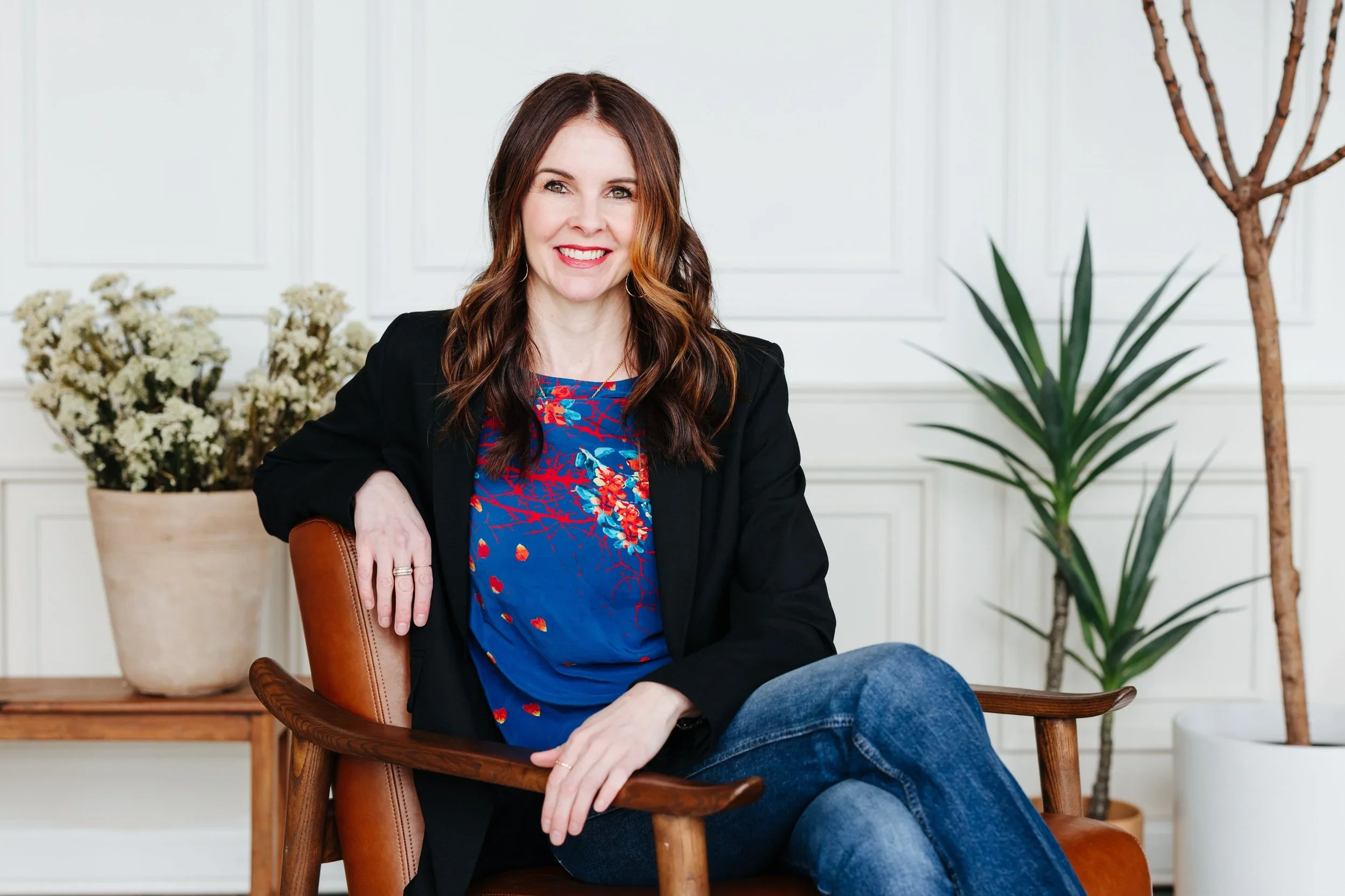 A woman with long brown hair wearing a black blazer and a blue floral top sitting on a brown chair, smiling, in a room with white walls, a large potted plant, and other decorative plants.