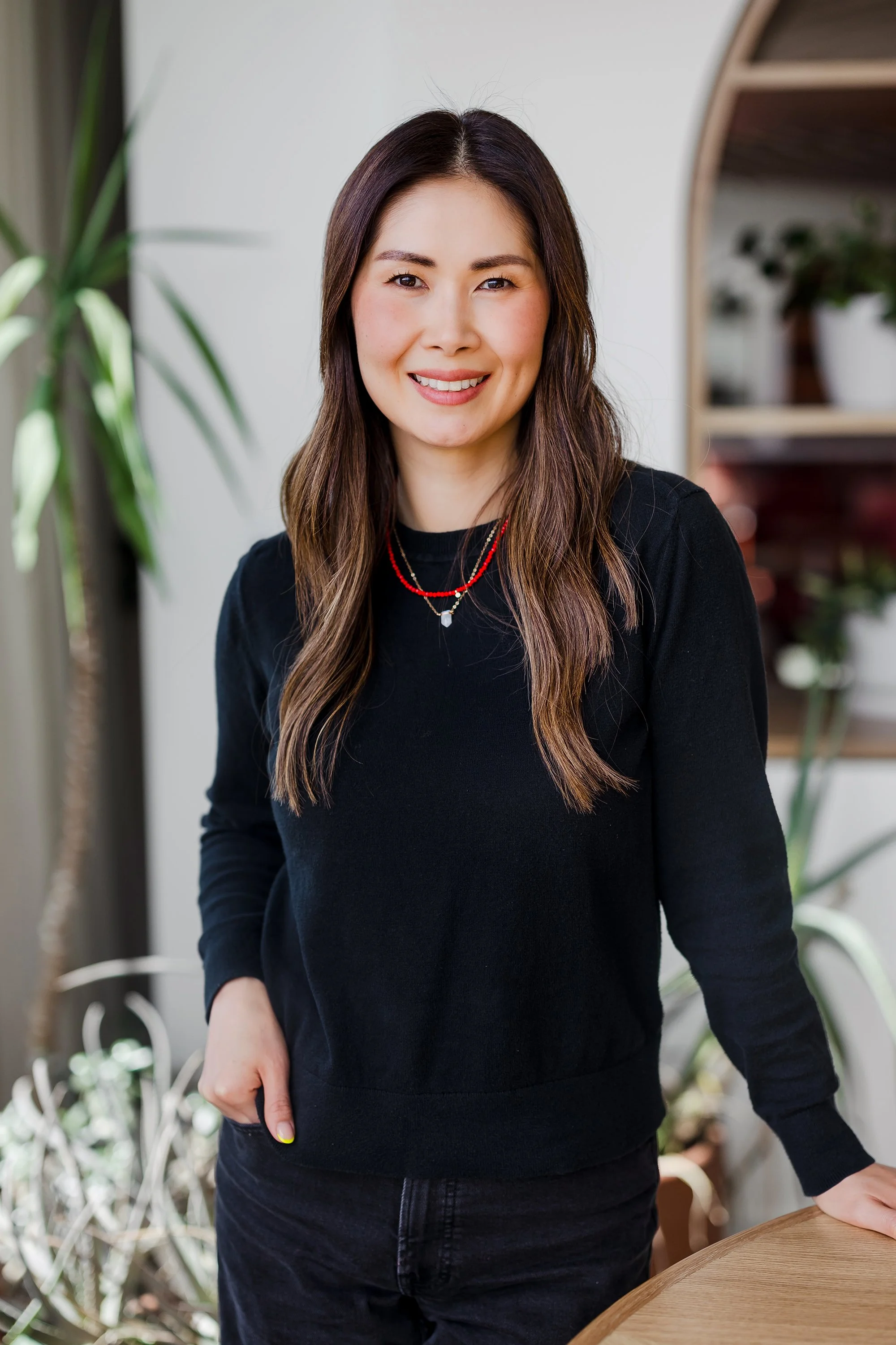 A woman with long brown hair, wearing a black top and red necklace, smiling while standing indoors near a round wooden table with plants in the background.
