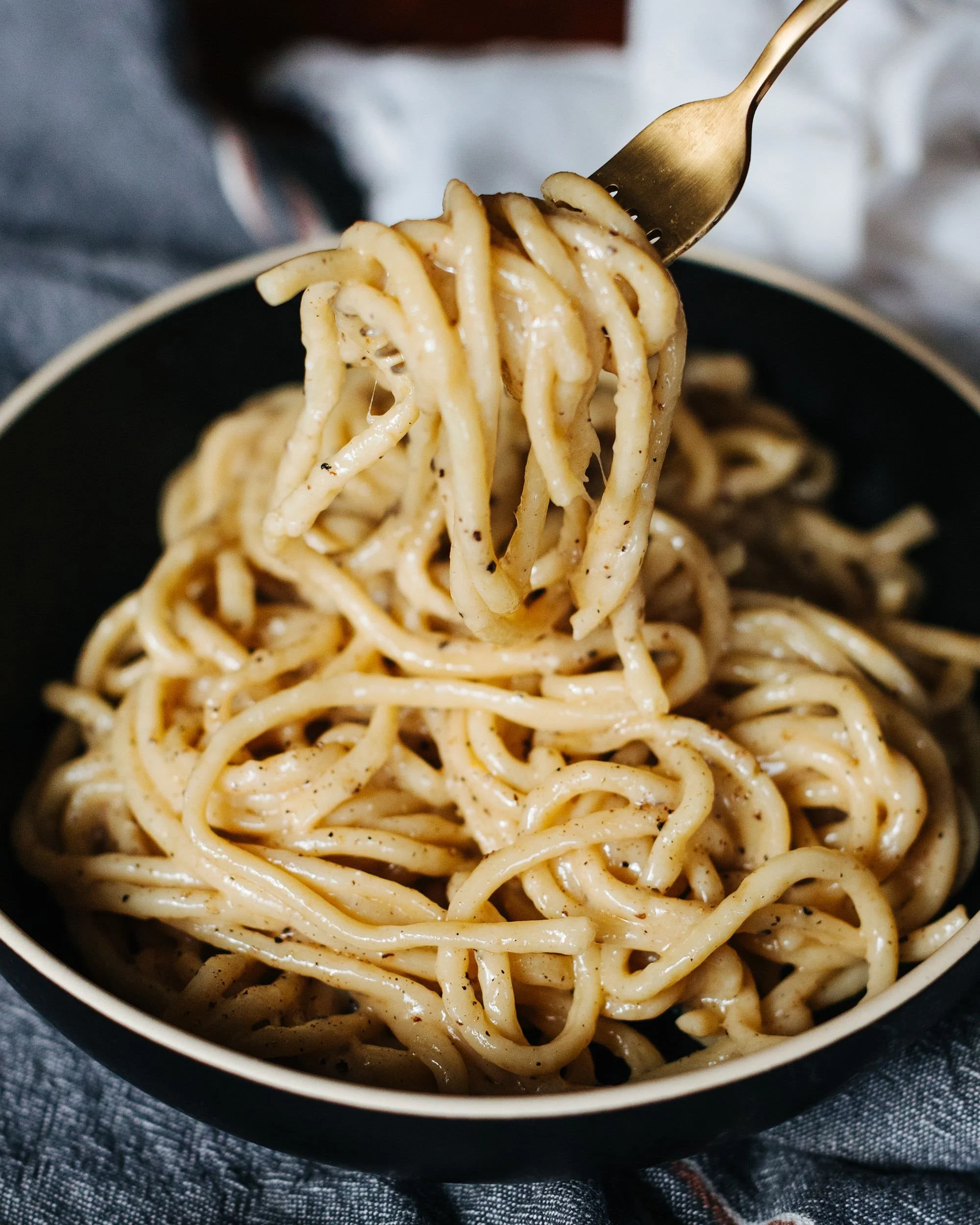 Close-up of cooked spaghetti with black pepper in a black bowl, with a fork lifting a portion of pasta, on a dark gray cloth background.