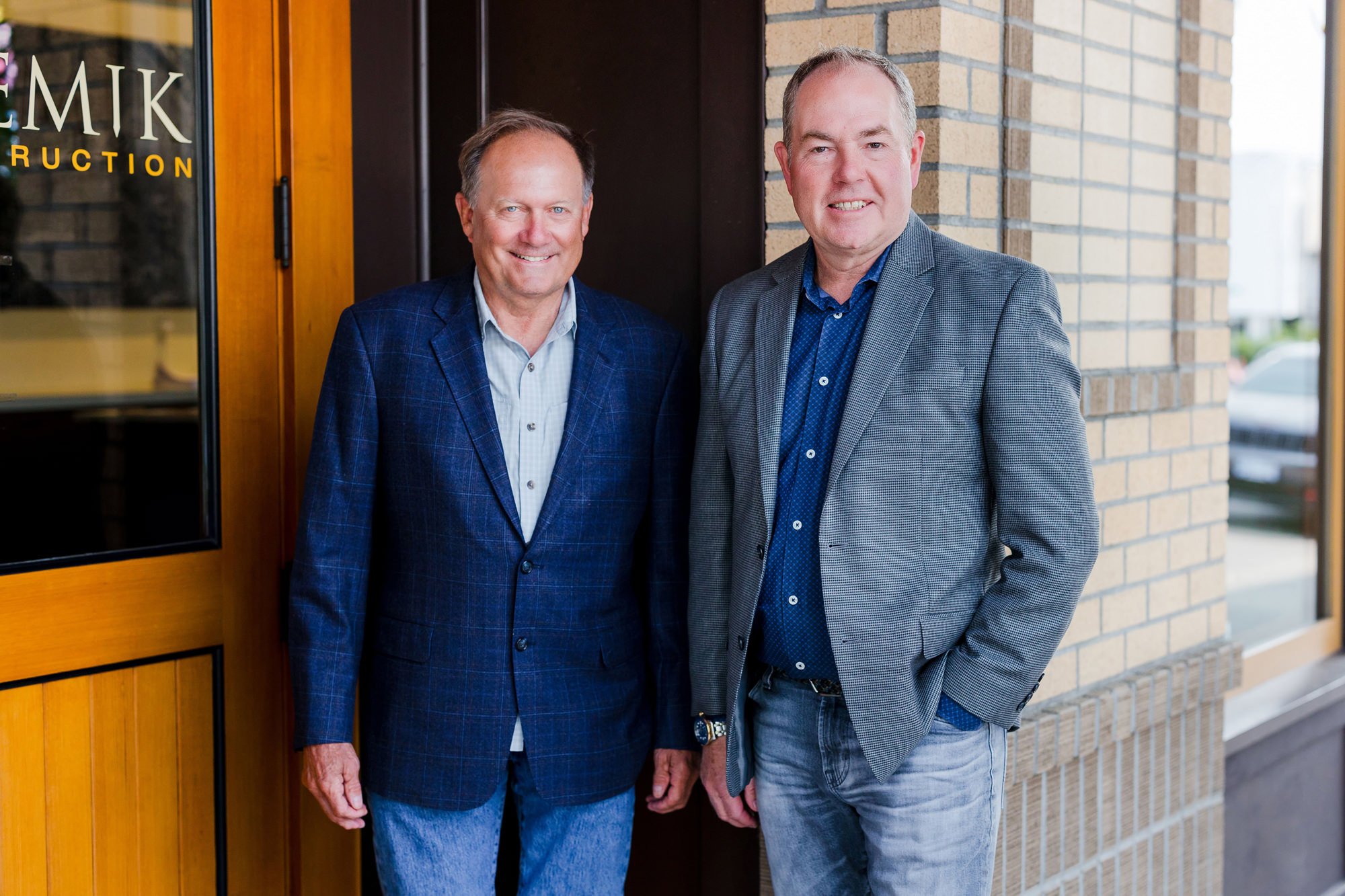 Two middle-aged men in suits standing outside an entrance with a glass door and brick wall, smiling at the camera.