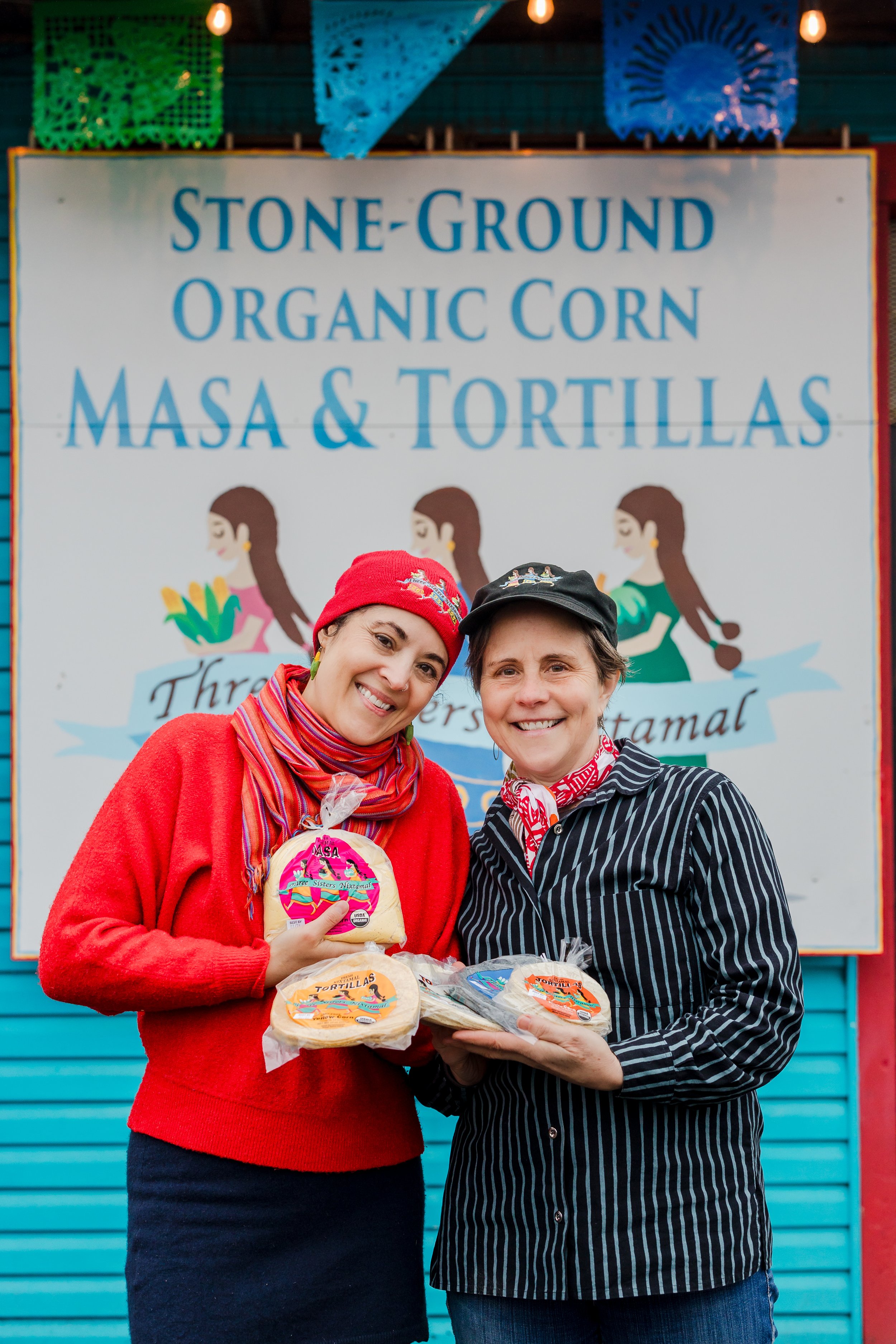 Two women smiling, holding bags of corn tortillas, standing in front of a sign for a tortilla stand.