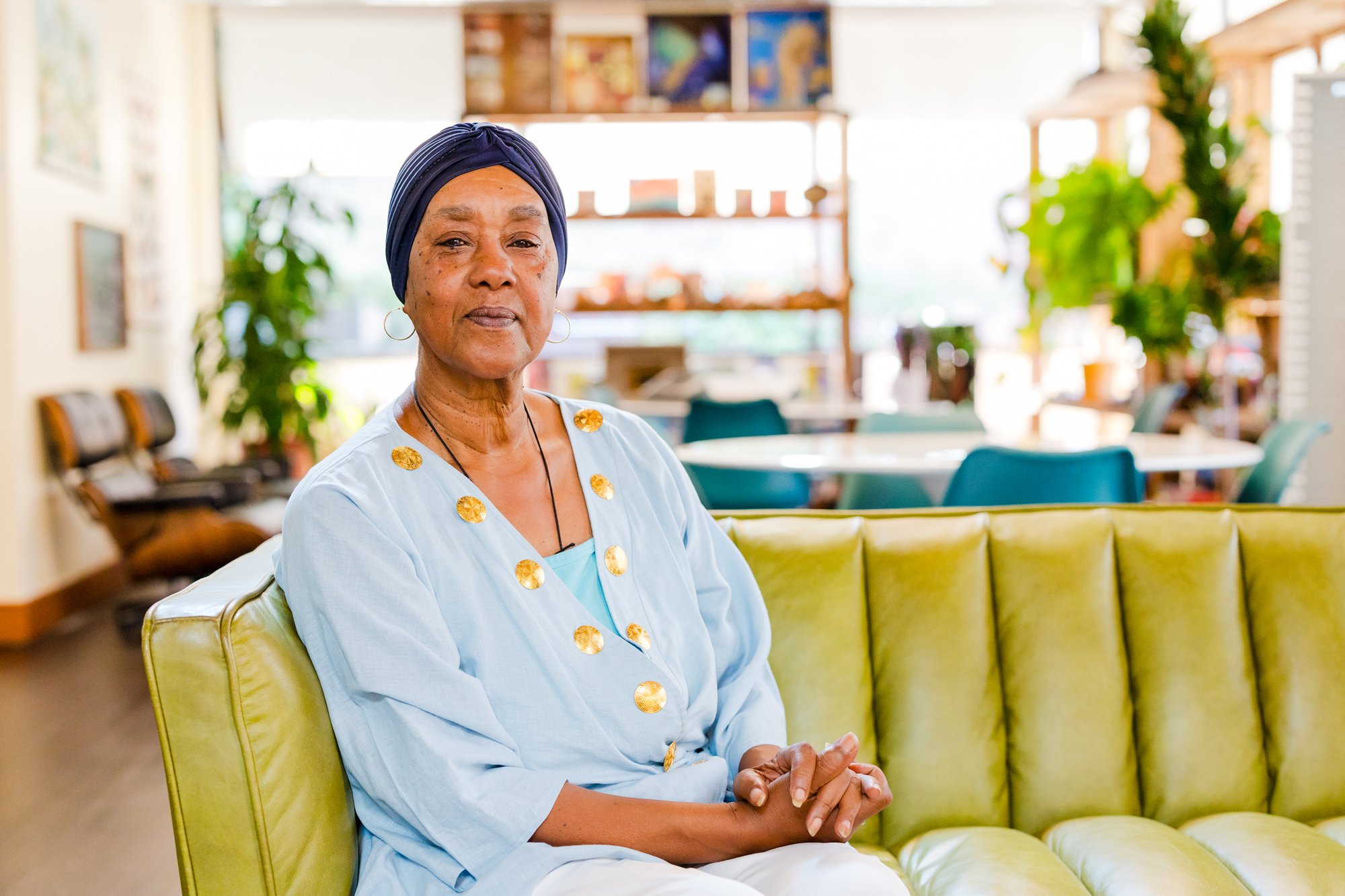 An elderly woman sitting on a yellow-green couch in a bright, modern space with plants and tables in the background.