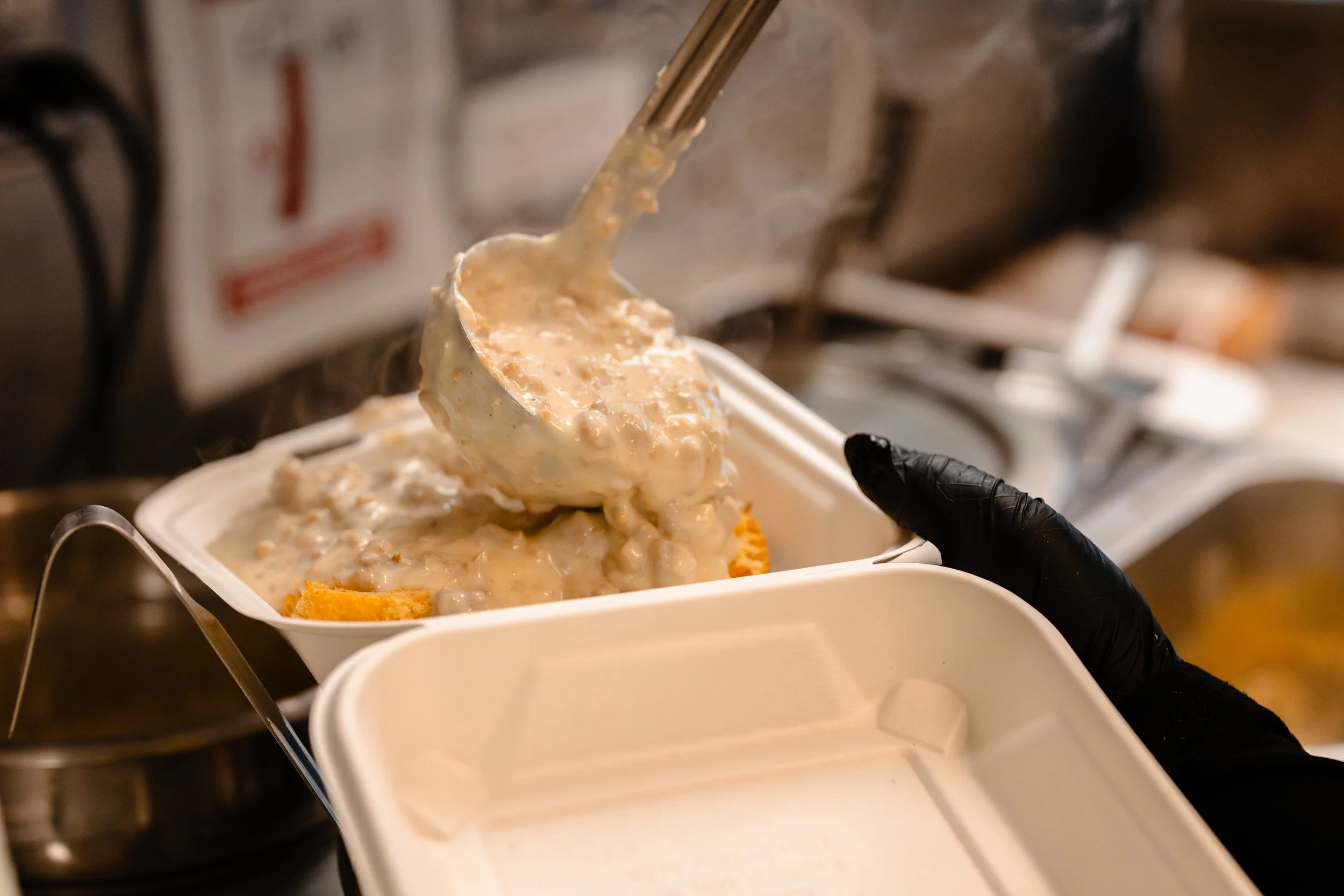 A person wearing black gloves serves a scoop of pot pie filling from a metal utensil into a foam takeout container.