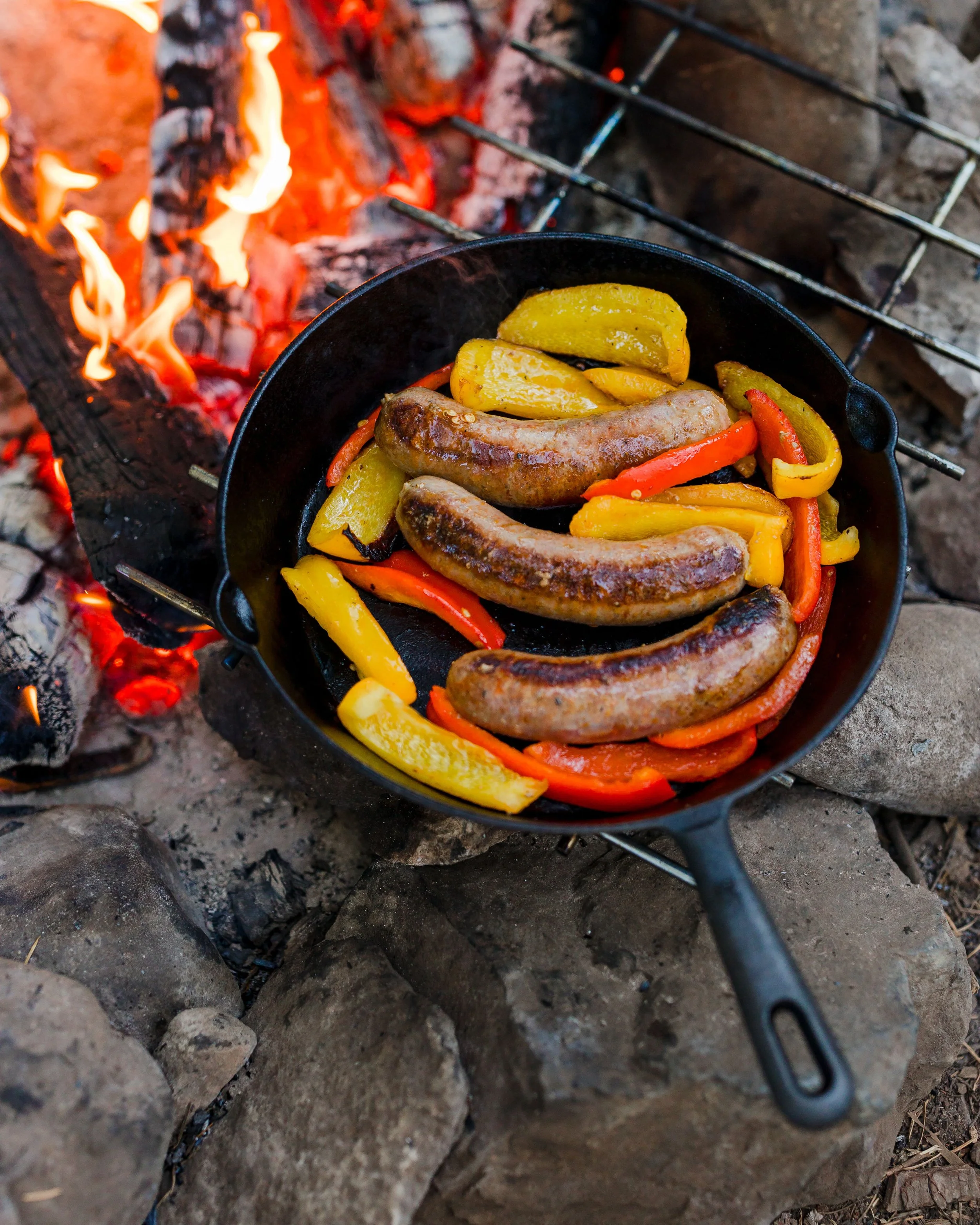 Cast iron skillet with grilled sausages, yellow and red bell peppers cooking over an open campfire surrounded by rocks.