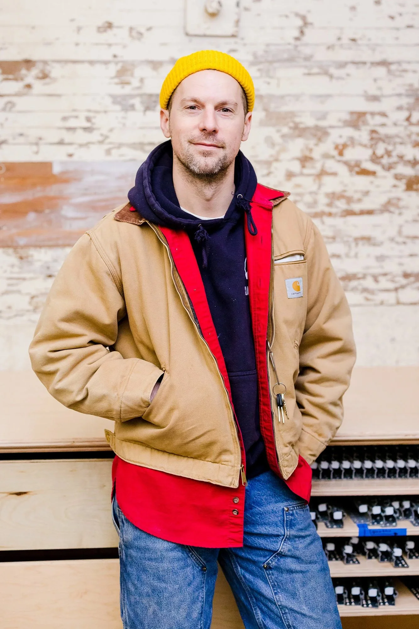 A man wearing a yellow beanie, navy hoodie, and tan Carhartt jacket stands in front of a rustic wooden wall.