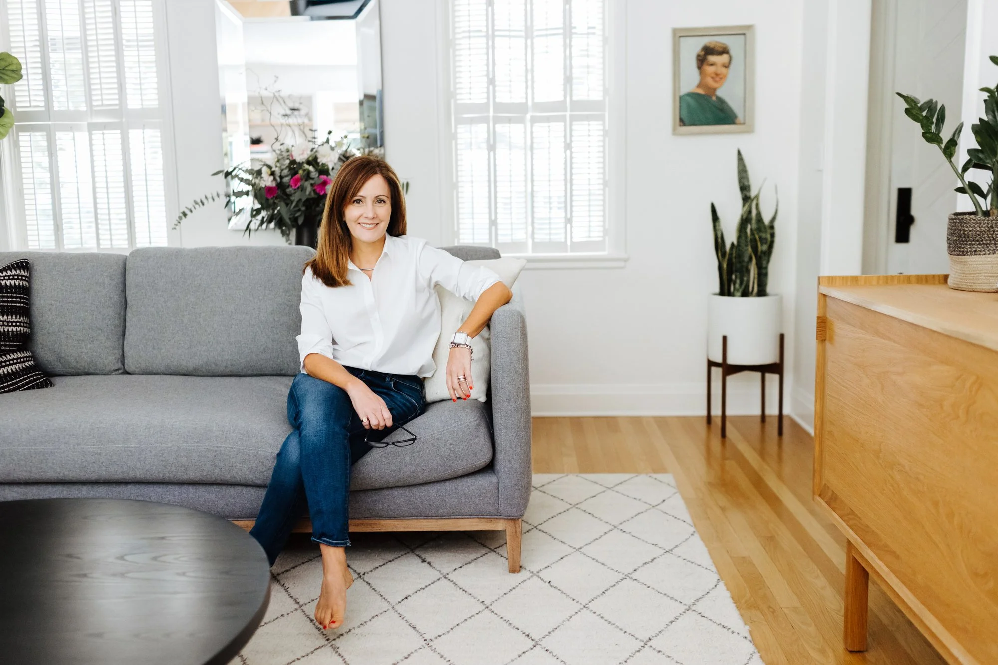 A woman with shoulder-length brown hair, wearing a white blouse and blue jeans, sitting on a gray sofa in a bright living room, smiling at the camera. There are windows with white blinds behind her, and the room is decorated with plants, a framed por