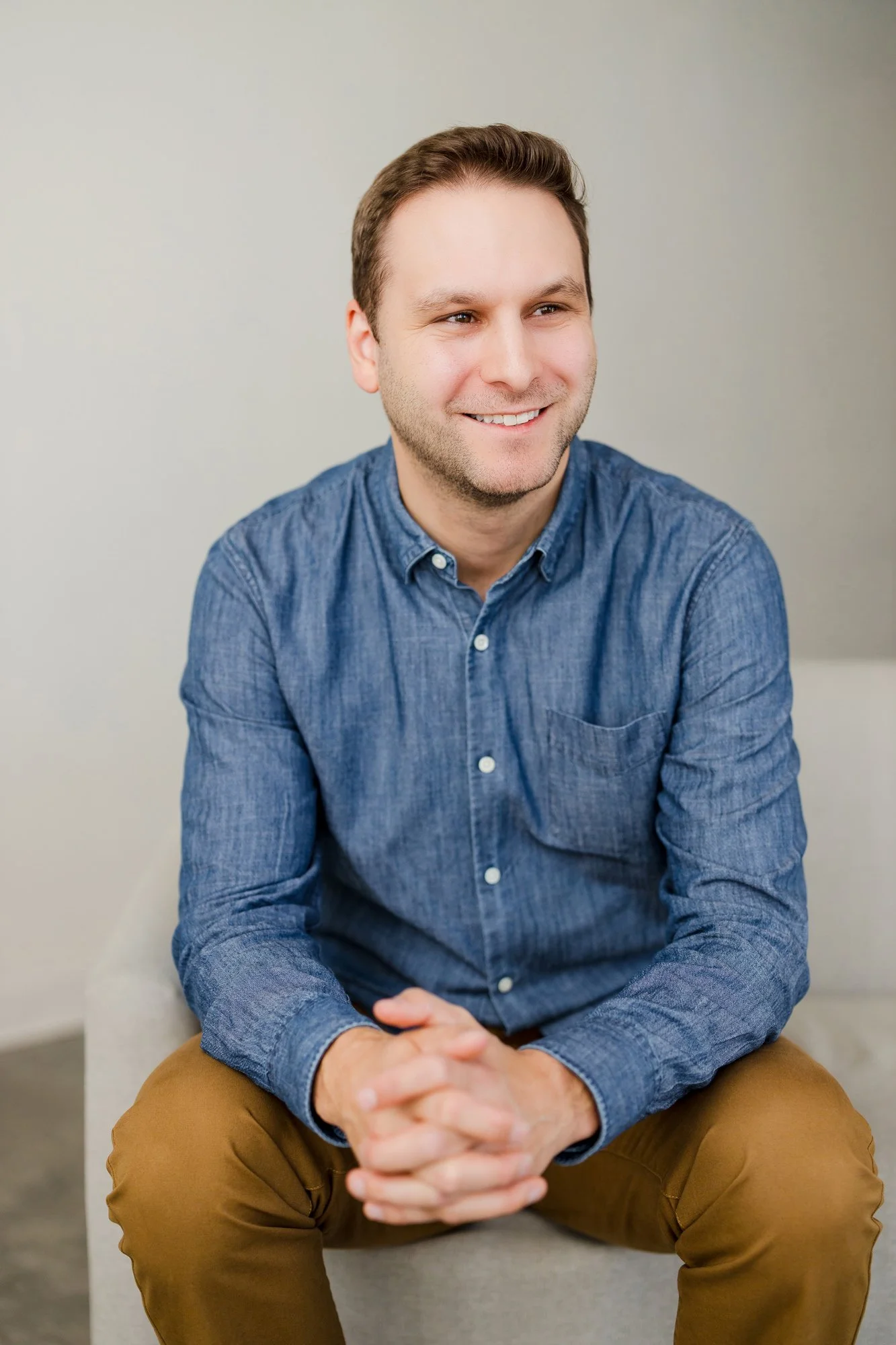 A smiling man with short brown hair wearing a blue button-up shirt and tan pants, sitting on a light-colored chair against a plain wall.