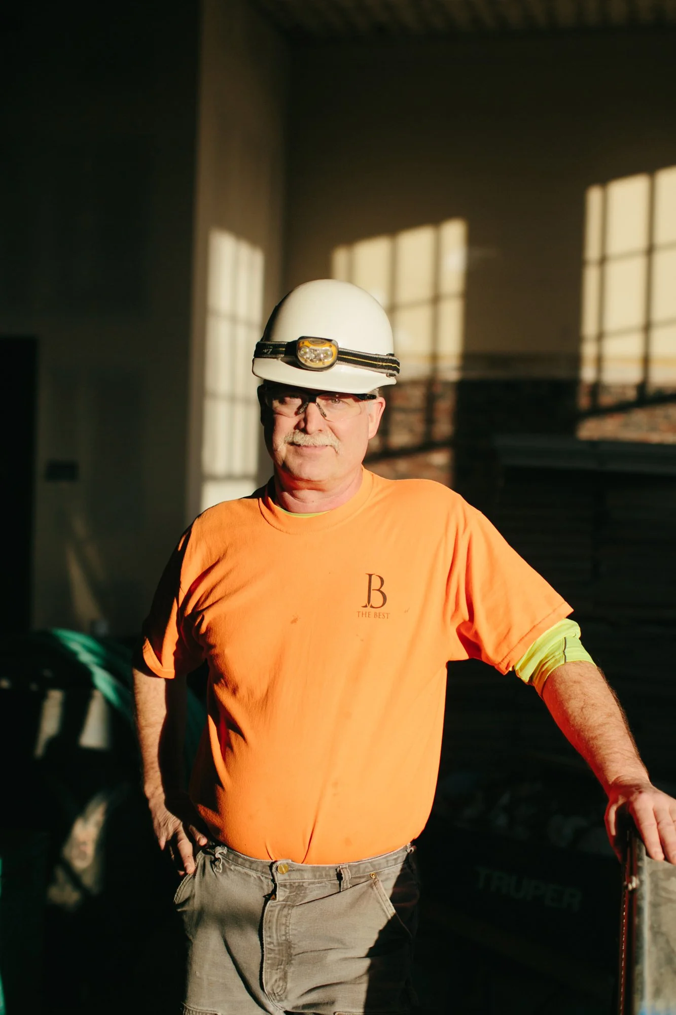 A construction worker wearing a white hard hat, safety glasses, and an orange shirt with a logo on it, standing in a sunlit room with windows in the background.
