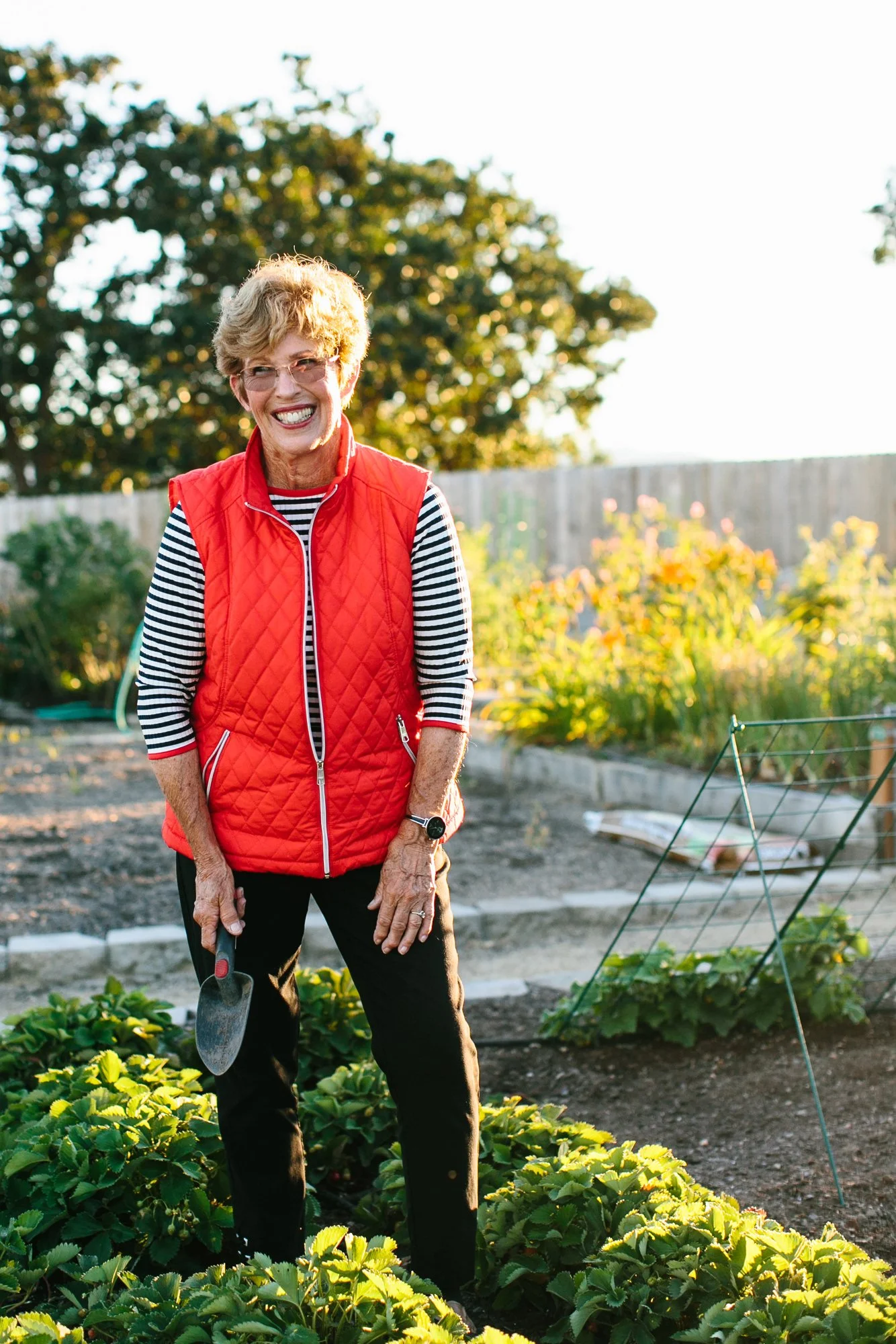 Happy elderly woman in garden holding a small shovel, wearing glasses, a red quilted vest over a striped shirt, and black pants, surrounded by green plants and flowers, with sunlight shining in the background.