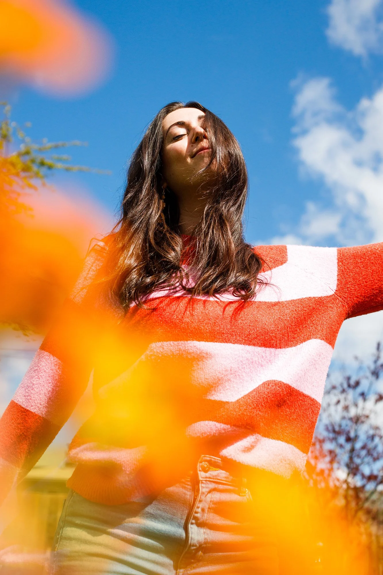 A woman standing outdoors with arms outstretched, wearing a red and white striped sweater, with her face tilted upward and eyes closed, under a blue sky with some clouds, and orange flowers in the foreground.