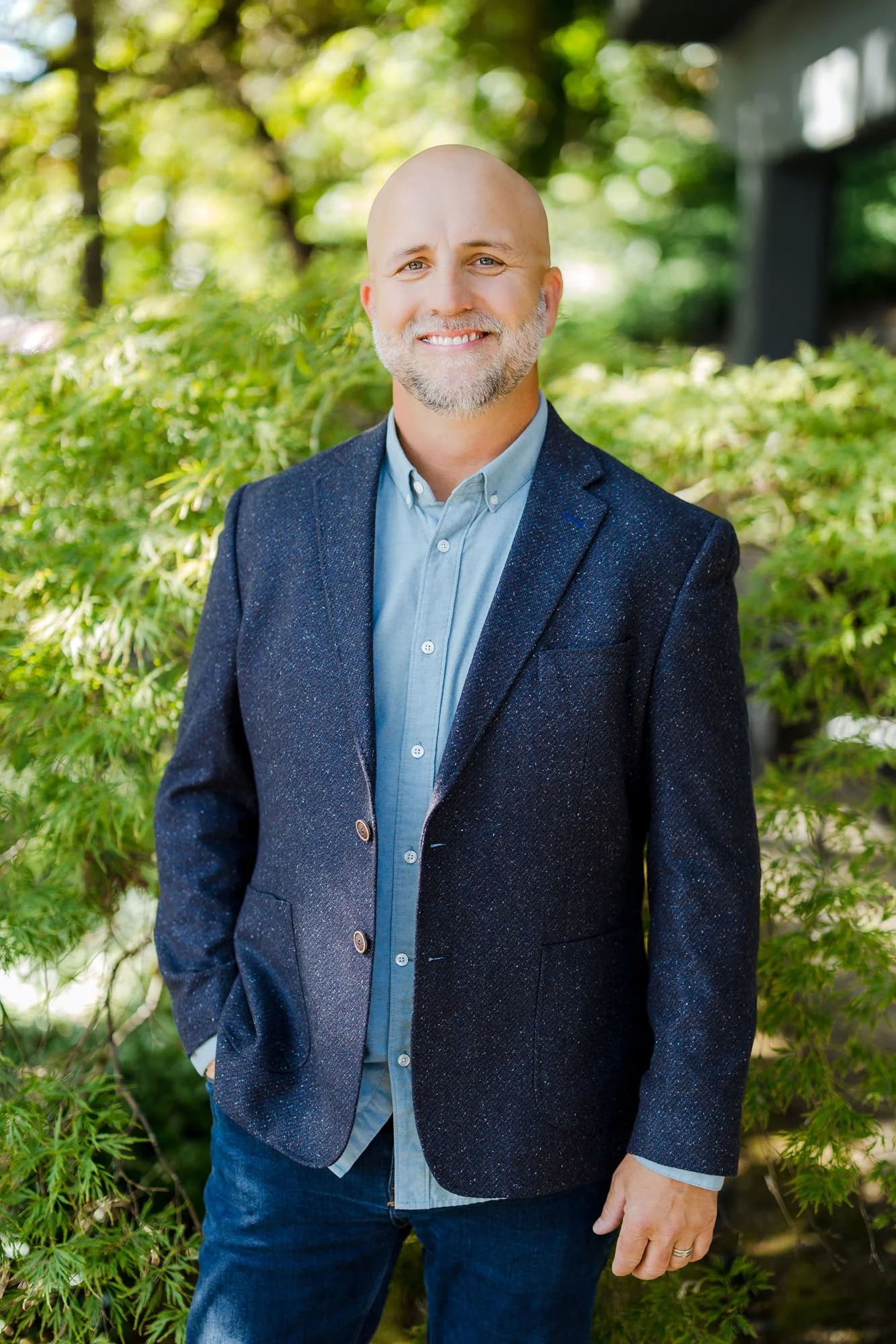 A man with a bald head, gray beard, and blue eyes, standing outdoors among green trees and bushes, smiling at the camera, wearing a navy blazer and light blue button-up shirt.
