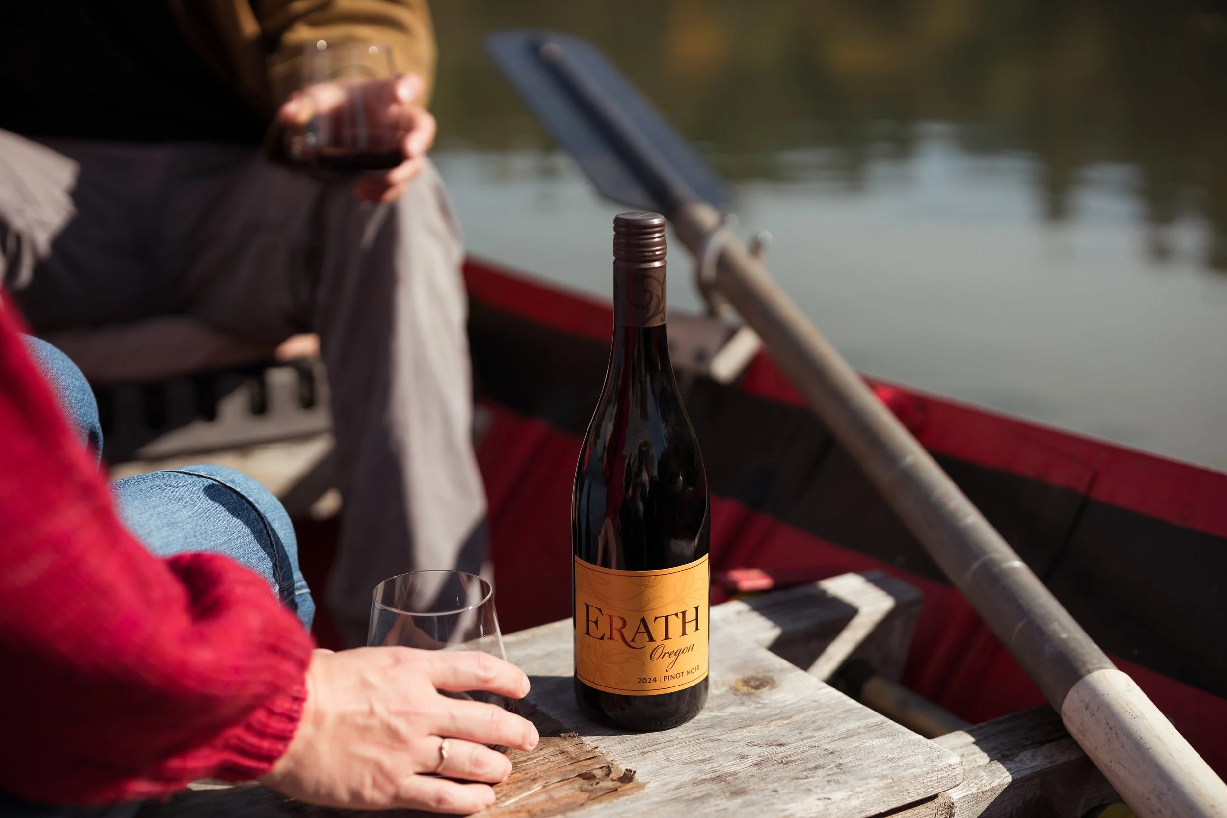 A close-up of a hand with a wedding ring holding a glass of red wine on a wooden surface next to a bottle of Erath Oregon Pinot Noir, with a boat and two people in the background on a body of water.