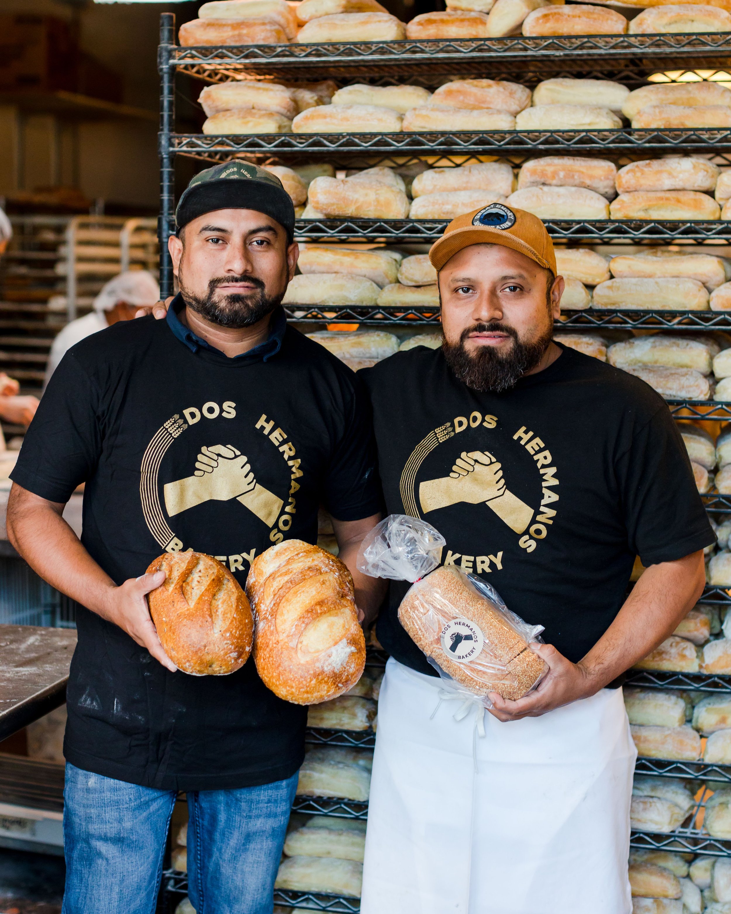 Two men wearing black 'Dos Hermanos Bakery' T-shirts holding loaves of bread, standing in front of a rack filled with bread in a bakery.