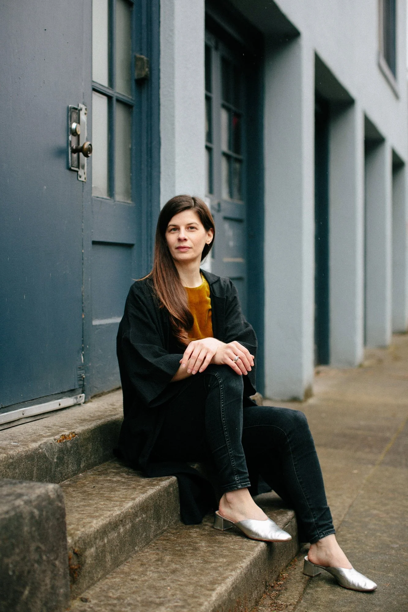 A woman with long brown hair sitting on concrete steps outside a building with gray walls and blue doors, wearing a black coat, black pants, and silver heels.