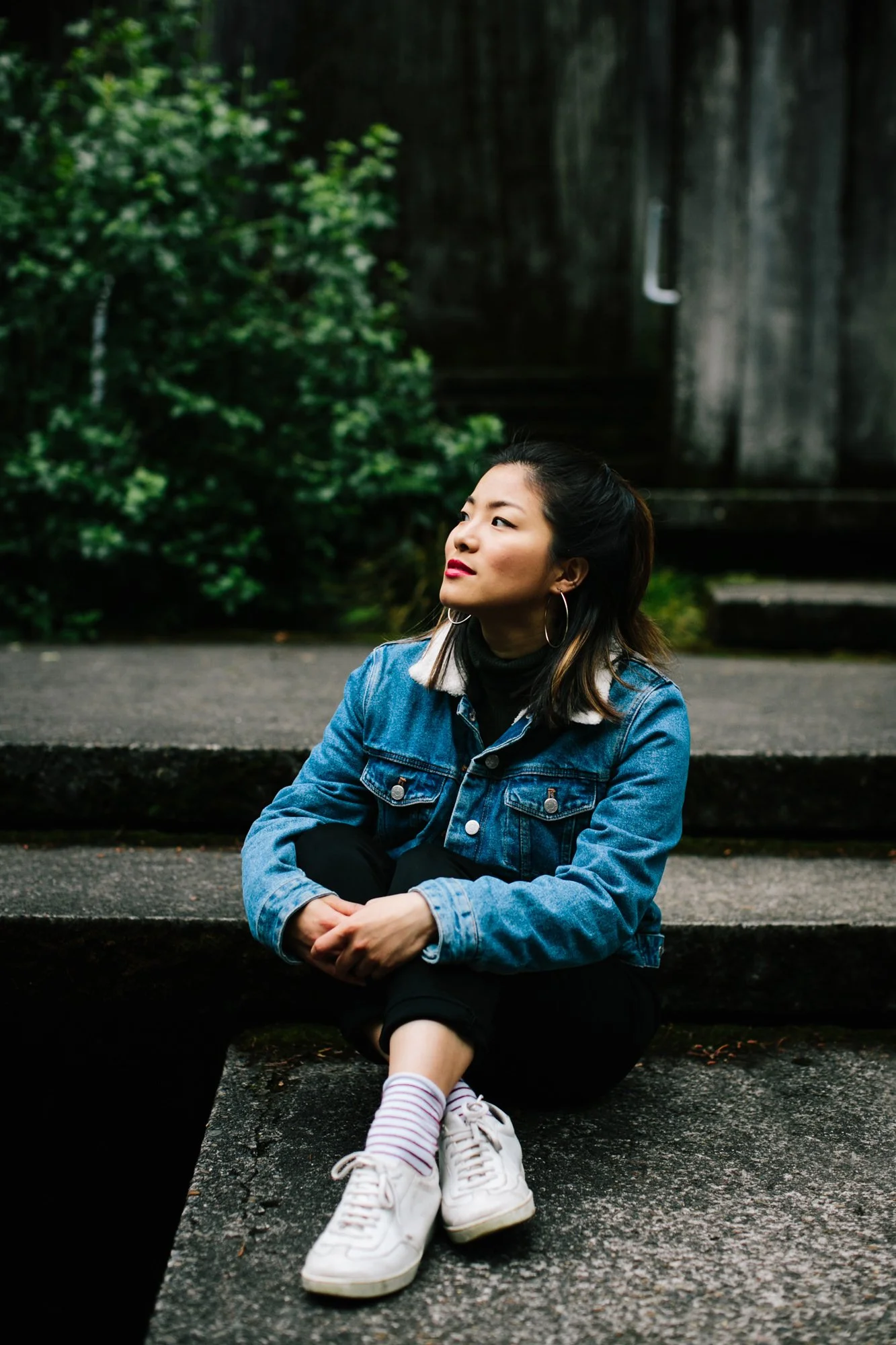 A young woman sitting on stone steps outdoors, wearing a denim jacket, black pants, white sneakers, and striped socks, looking to the side with a thoughtful expression, with greenery and a wooden structure in the background.