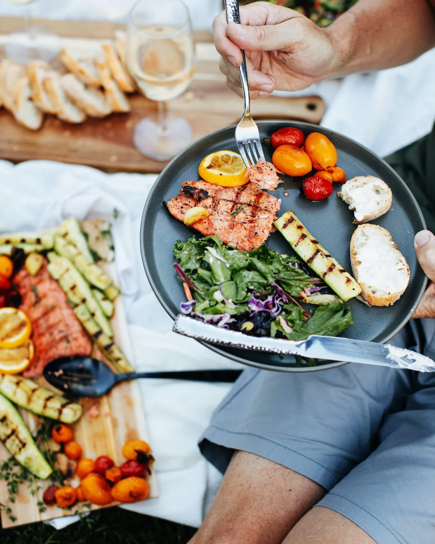 A person holding a black plate with grilled vegetables, salad, grilled fish, bread, and cherry tomatoes at an outdoor meal, with wine and additional food on a wooden table in the background.