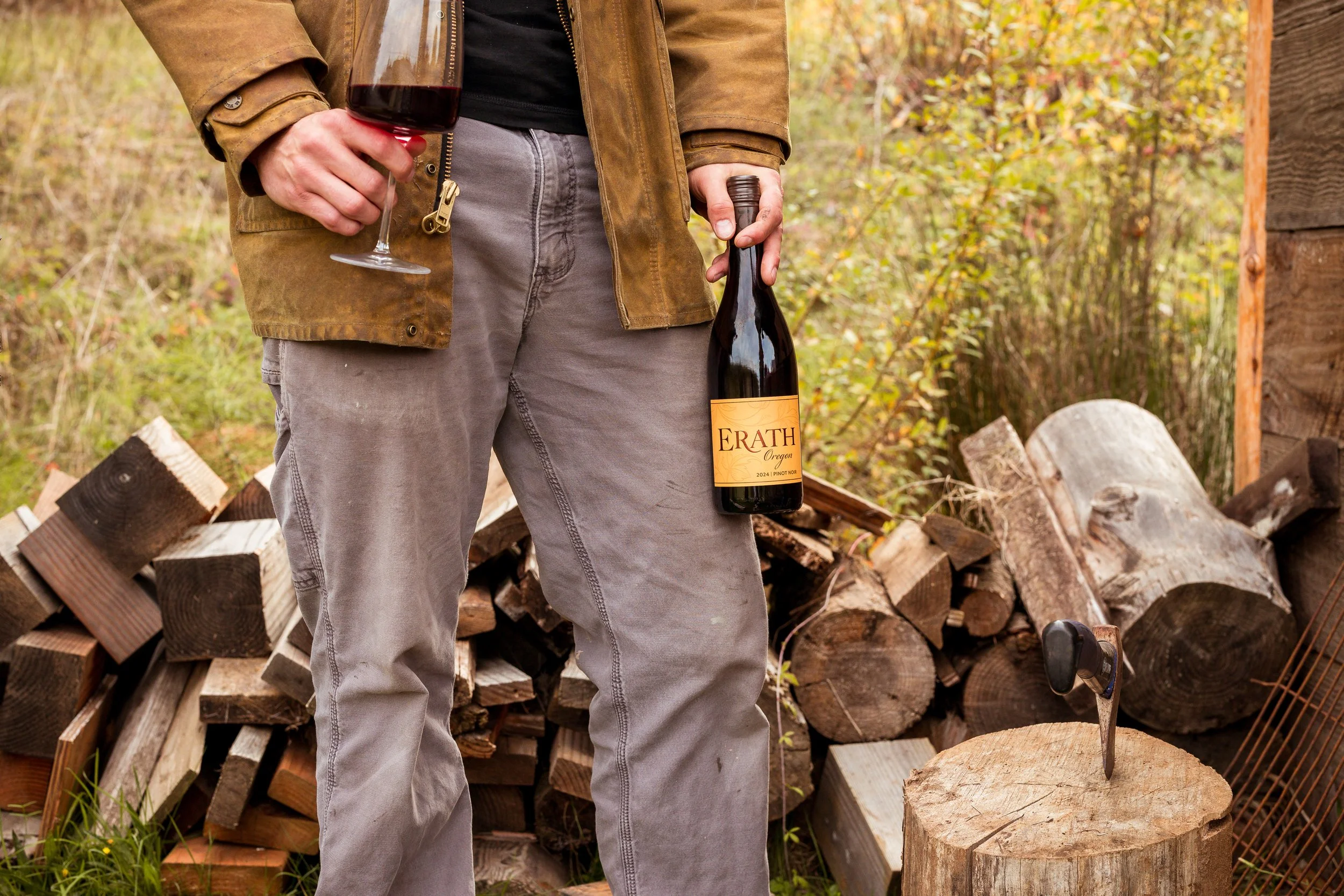A person holding a glass of red wine and a bottle of wine outdoors, with firewood stacked in the background.