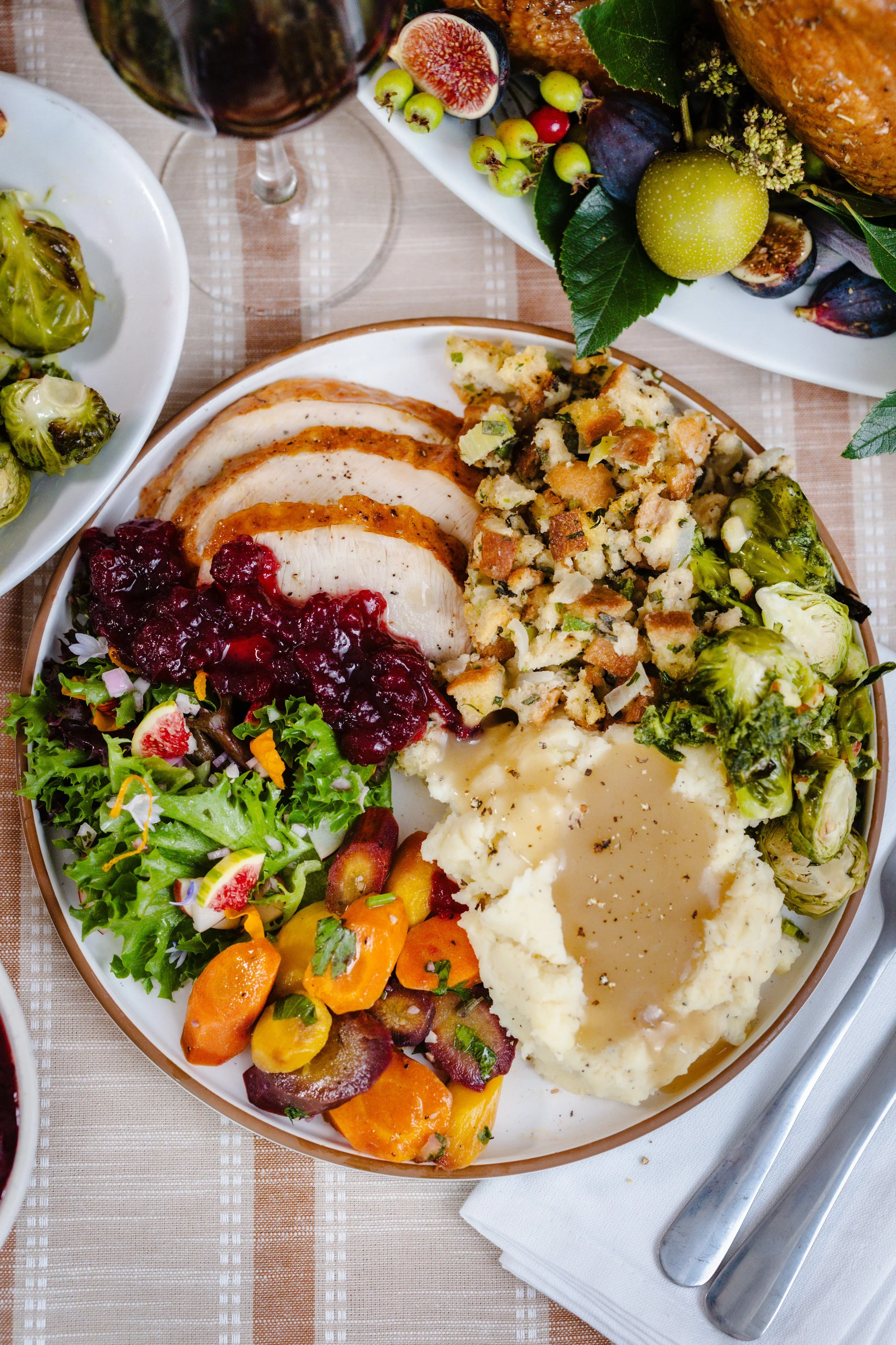 Plate of Thanksgiving foods including sliced turkey with cranberry sauce, mashed potatoes with gravy, stuffing, green beans, sweet potatoes, and a mixed green salad, next to a glass of red wine and a bowl of Brussels sprouts, on a checked tablecloth.