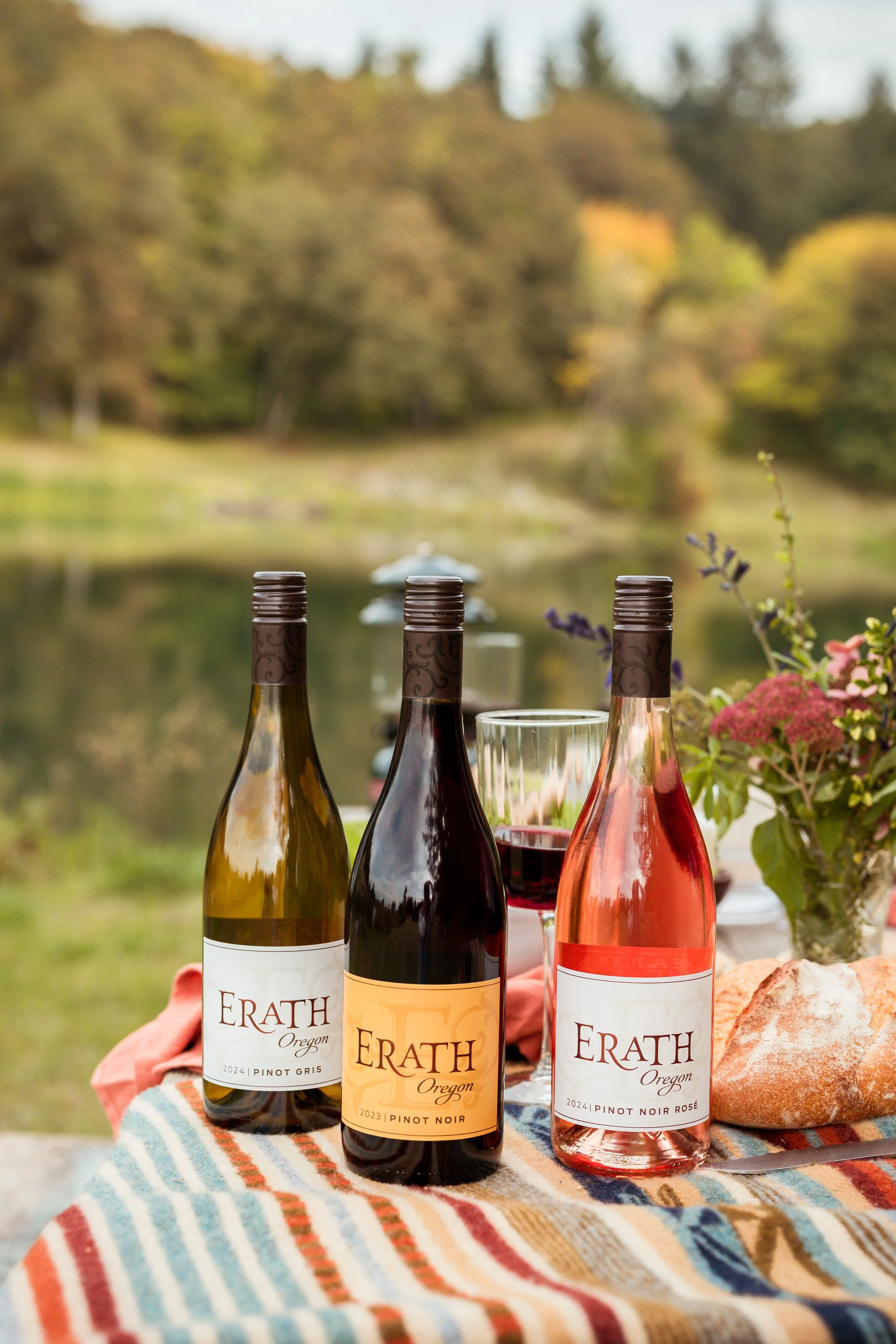 Three bottles of Erath Oregon wine, including Pinot Gris, Pinot Noir, and Pinot Noir Rosé, are set on a colorful striped cloth with a glass of red wine, a loaf of bread, and a vase of flowers, outdoors with a blurred lake and trees in the background.