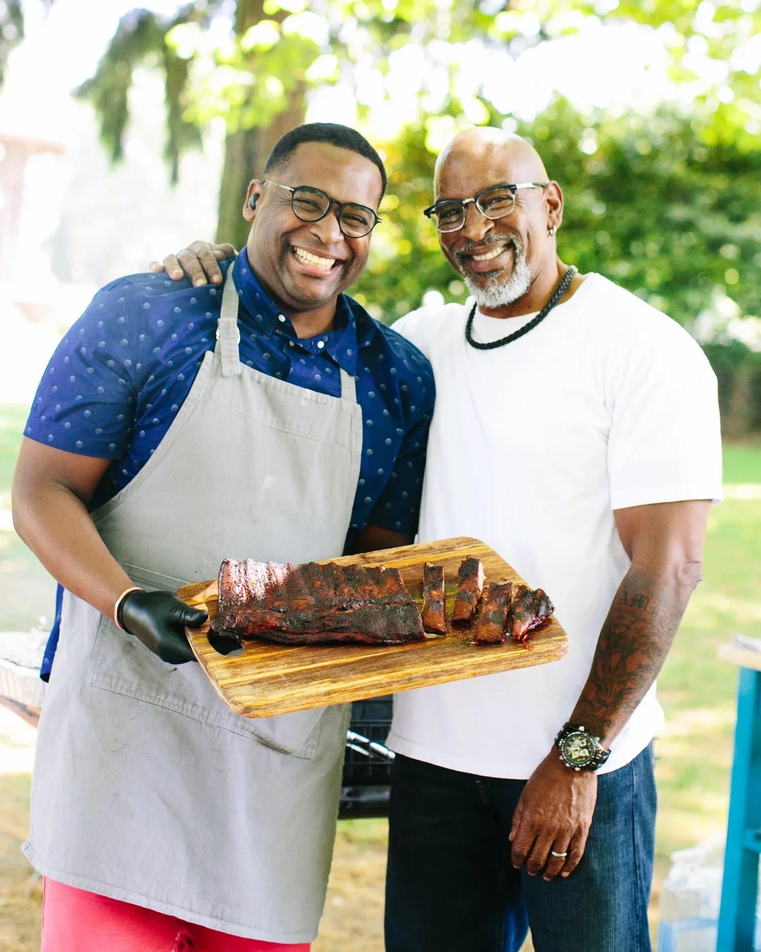 Two smiling men outdoors, one holding a wooden board with sliced barbecue ribs, the other with one arm around the first man.