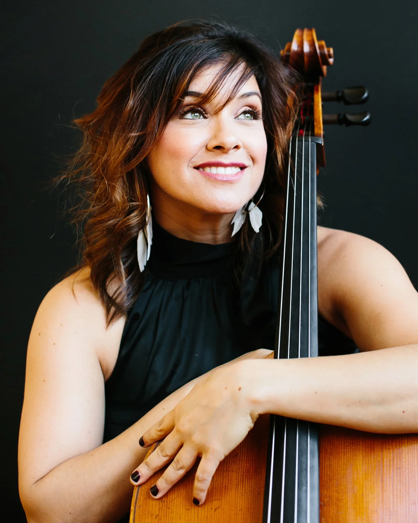 A woman with wavy brown hair and hoop earrings smiling and holding a cello against a dark background.