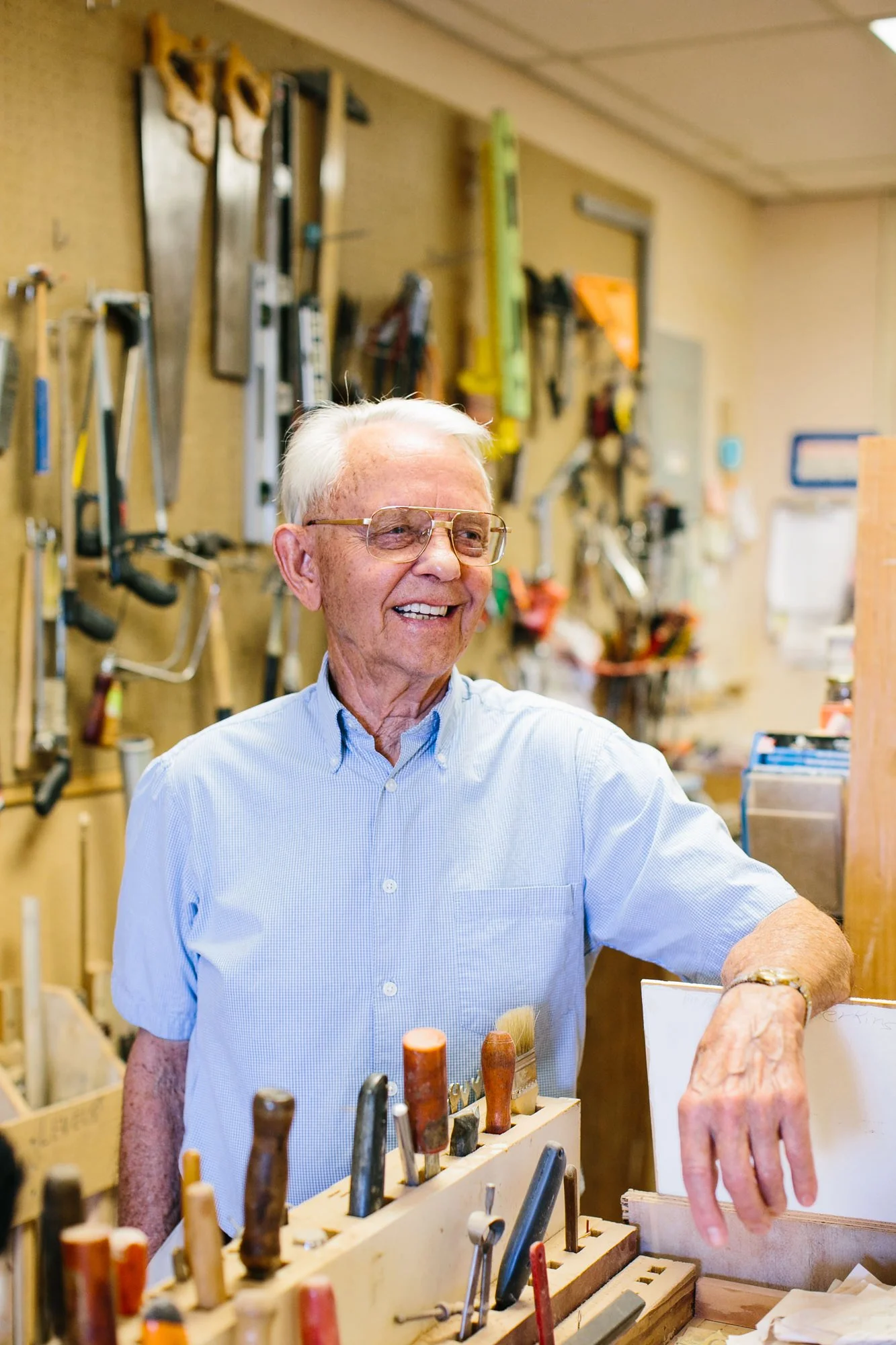 An elderly man smiling in a woodworking shop with various tools hanging on the wall behind him.