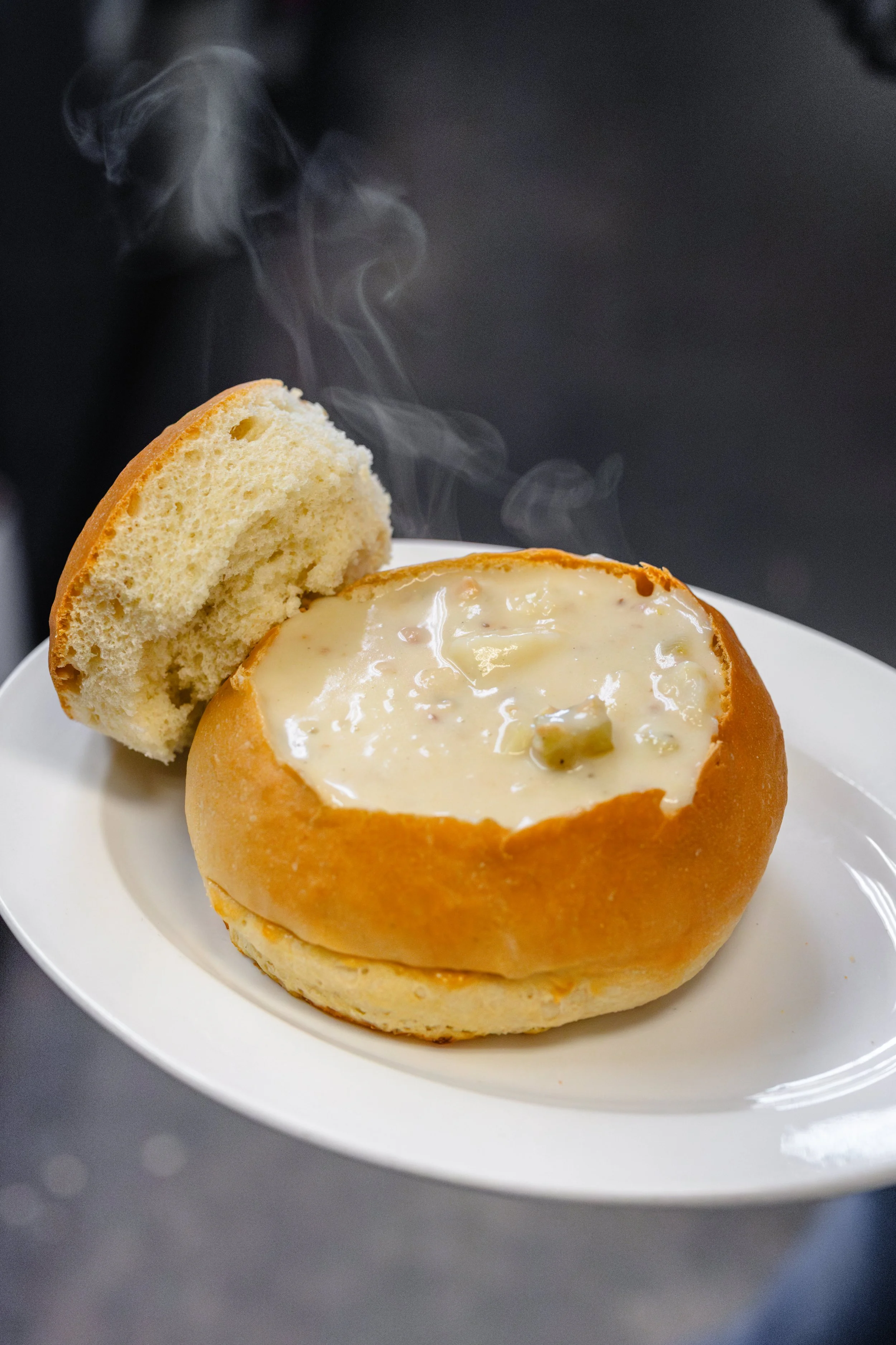 A bread bowl filled with creamy soup, placed on a white plate, with a piece of bread on the side, and steam rising from the hot soup.