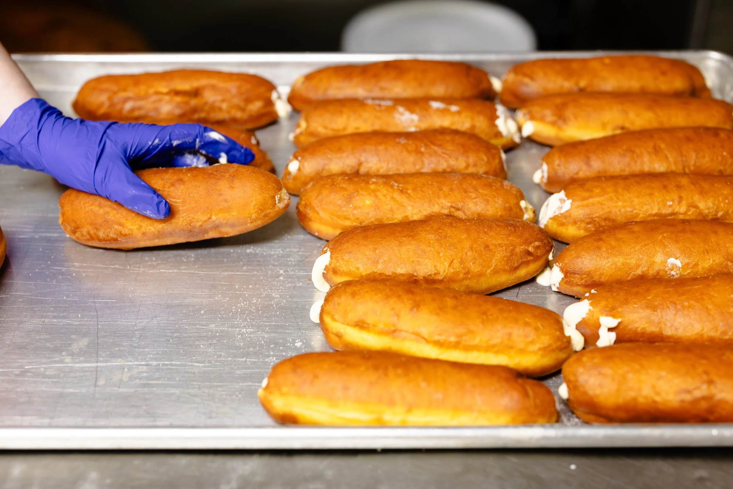 A person wearing a blue glove is placing a fried pastry filled with cream on a metal tray, with more fried pastries lined up on the tray.