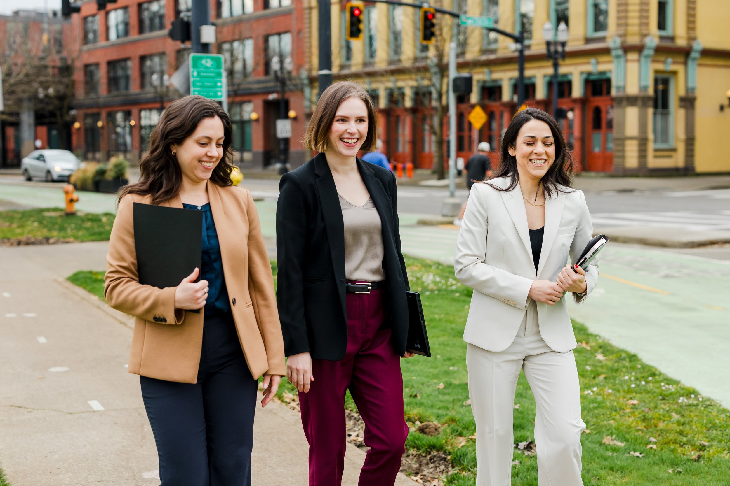 Three women walking outdoors in an urban area, smiling and holding folders or notebooks, dressed in business attire.