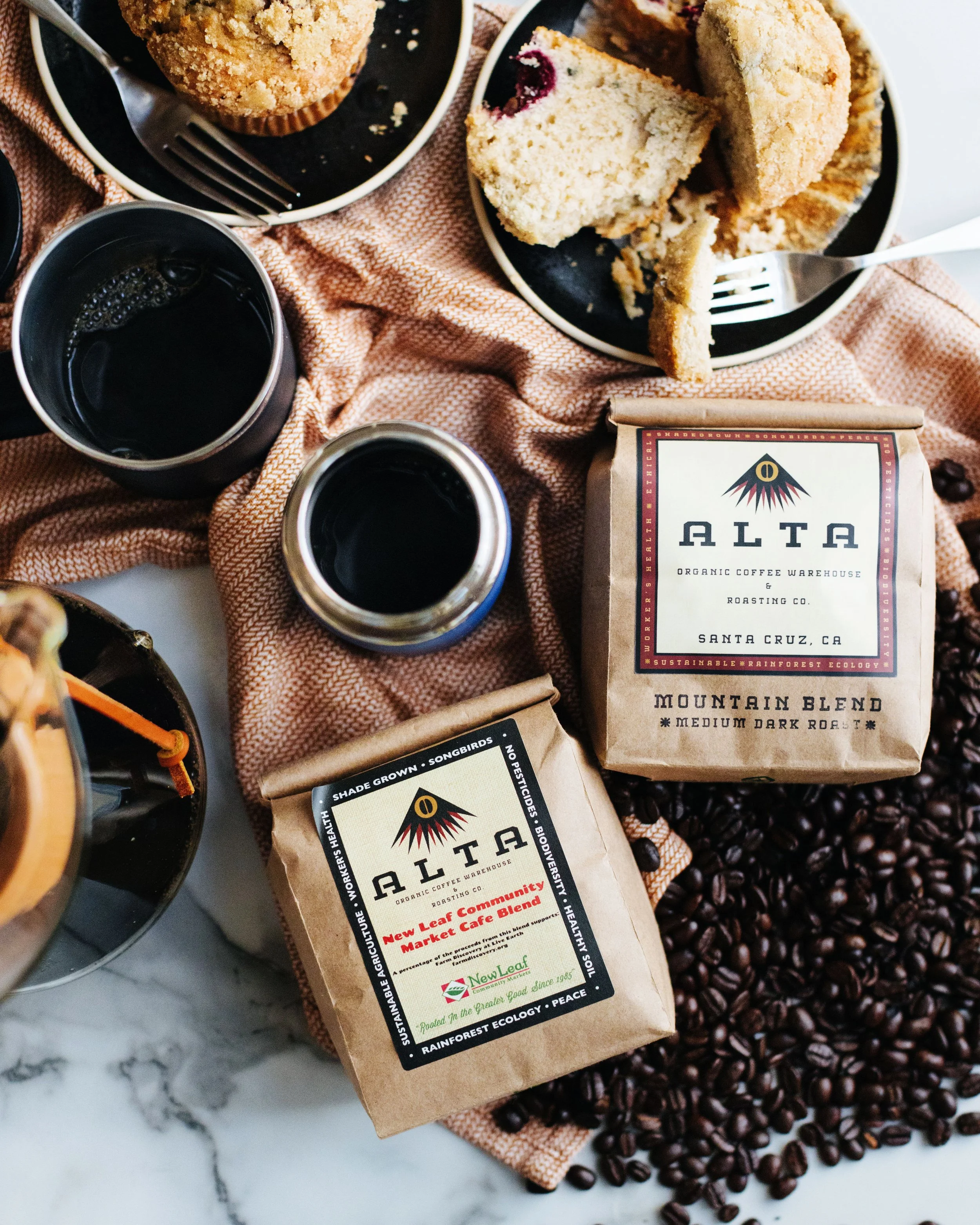Coffee beans, two bags of ALTA coffee, a cup of black coffee, a jar of black coffee, and sliced cake on a pink cloth.